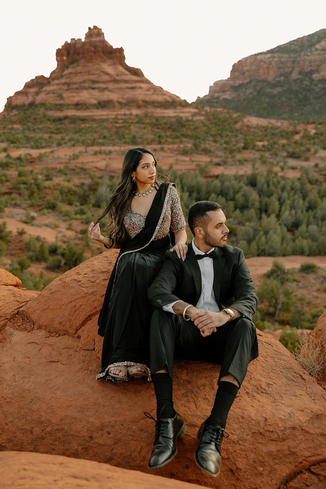 Dressed in formal attire, the couple poses on warm red rocks with Bell Rock stretching behind them.
