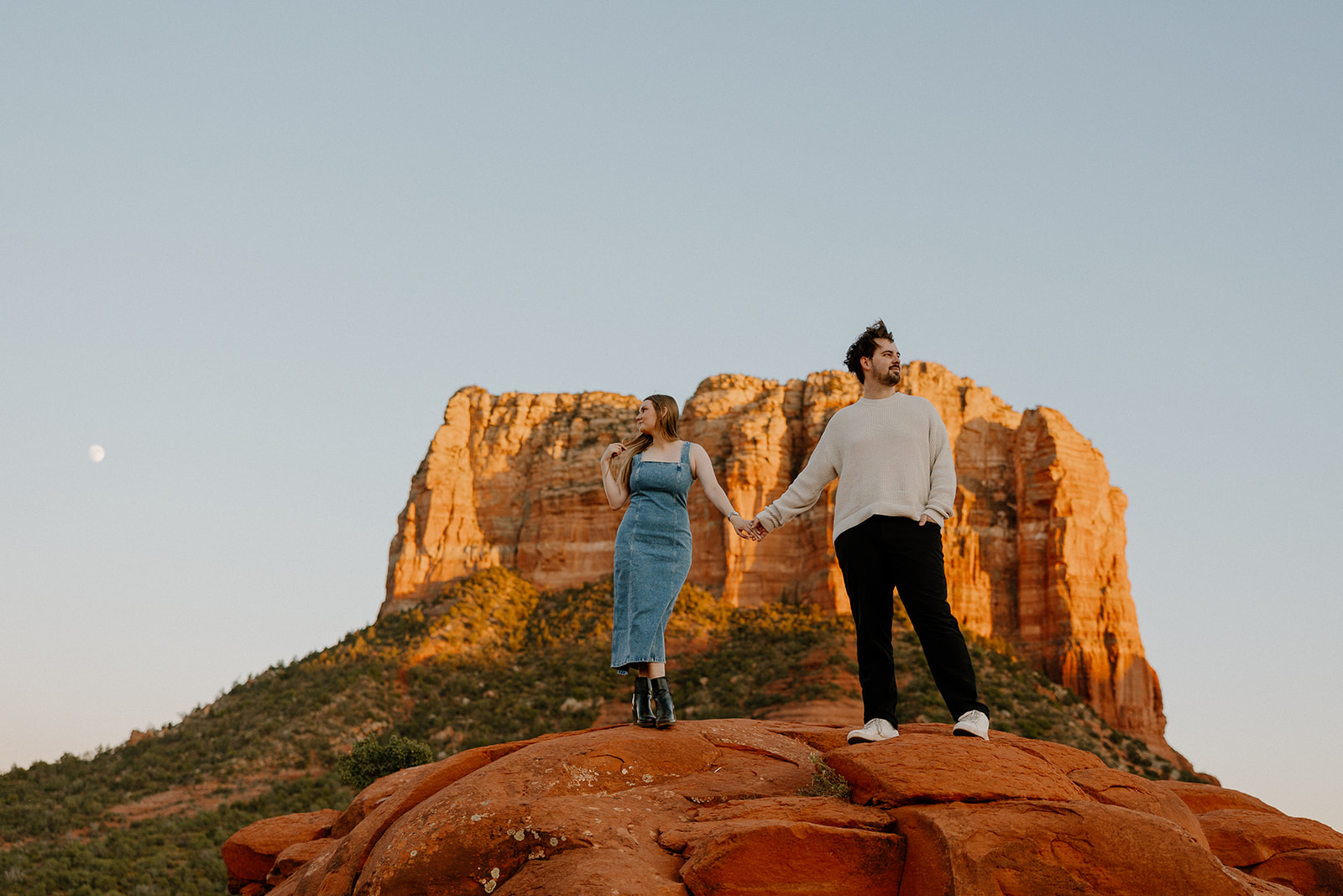 Woman wearing denim dress holding hands with man dressed casually standing on top of red rocks in Sedona