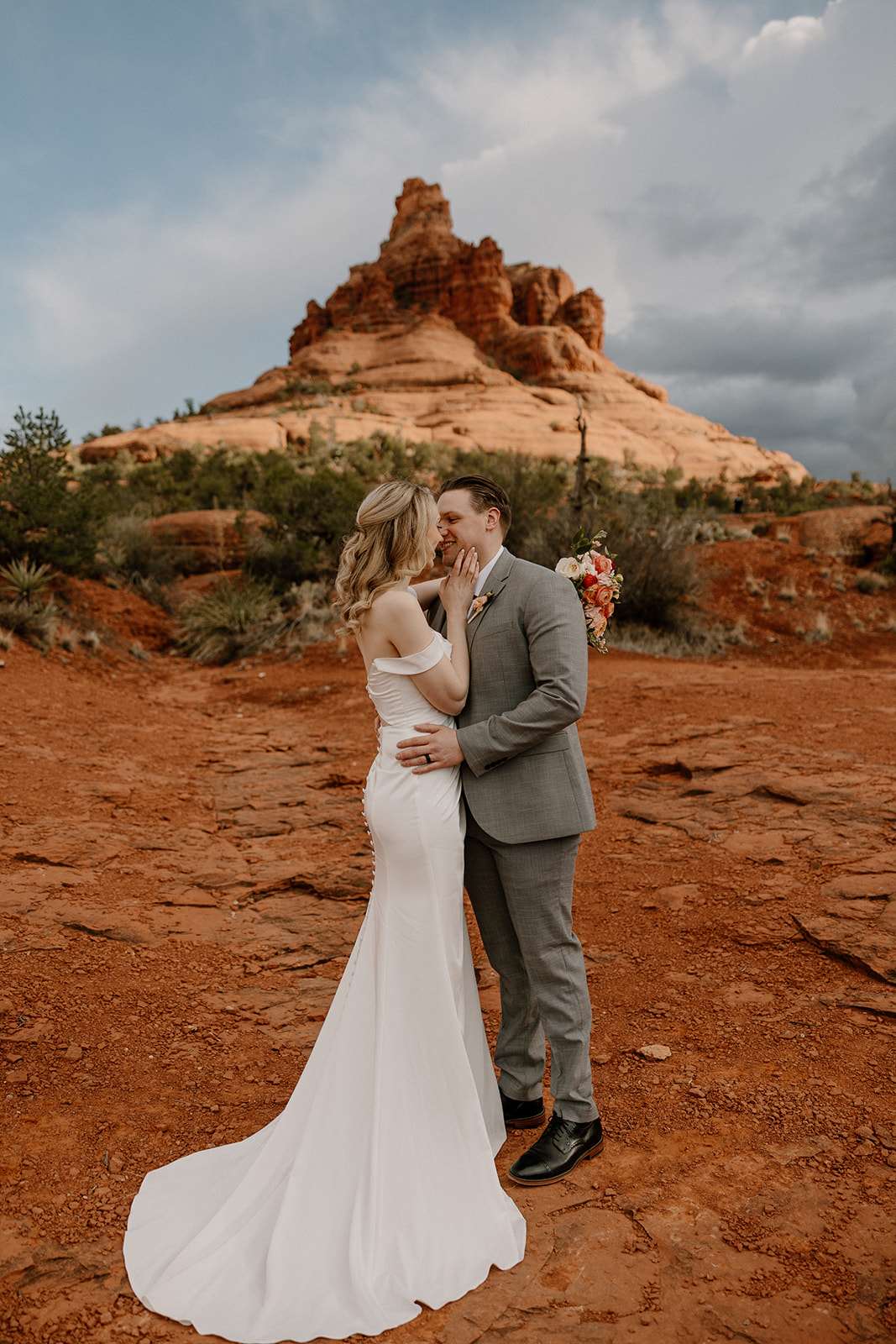 A bride and groom embrace in front of Bell Rock, her sleek white dress glowing against the red dirt backdrop.