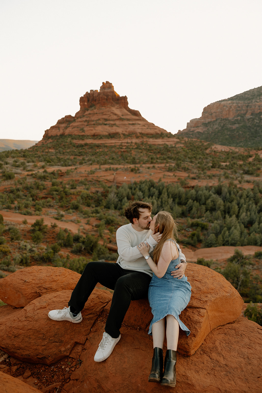 Sitting on a ledge at golden hour, the couple shares a kiss with Bell Rock rising in the distance.