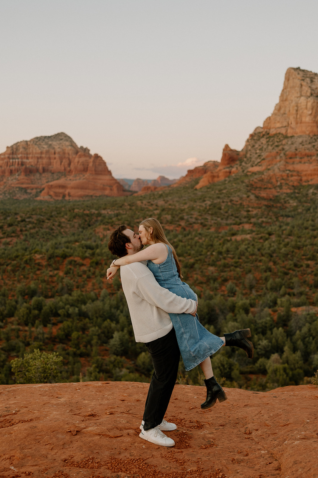 She wraps her legs around him mid-lift, laughing as he holds her close against a red rock backdrop.