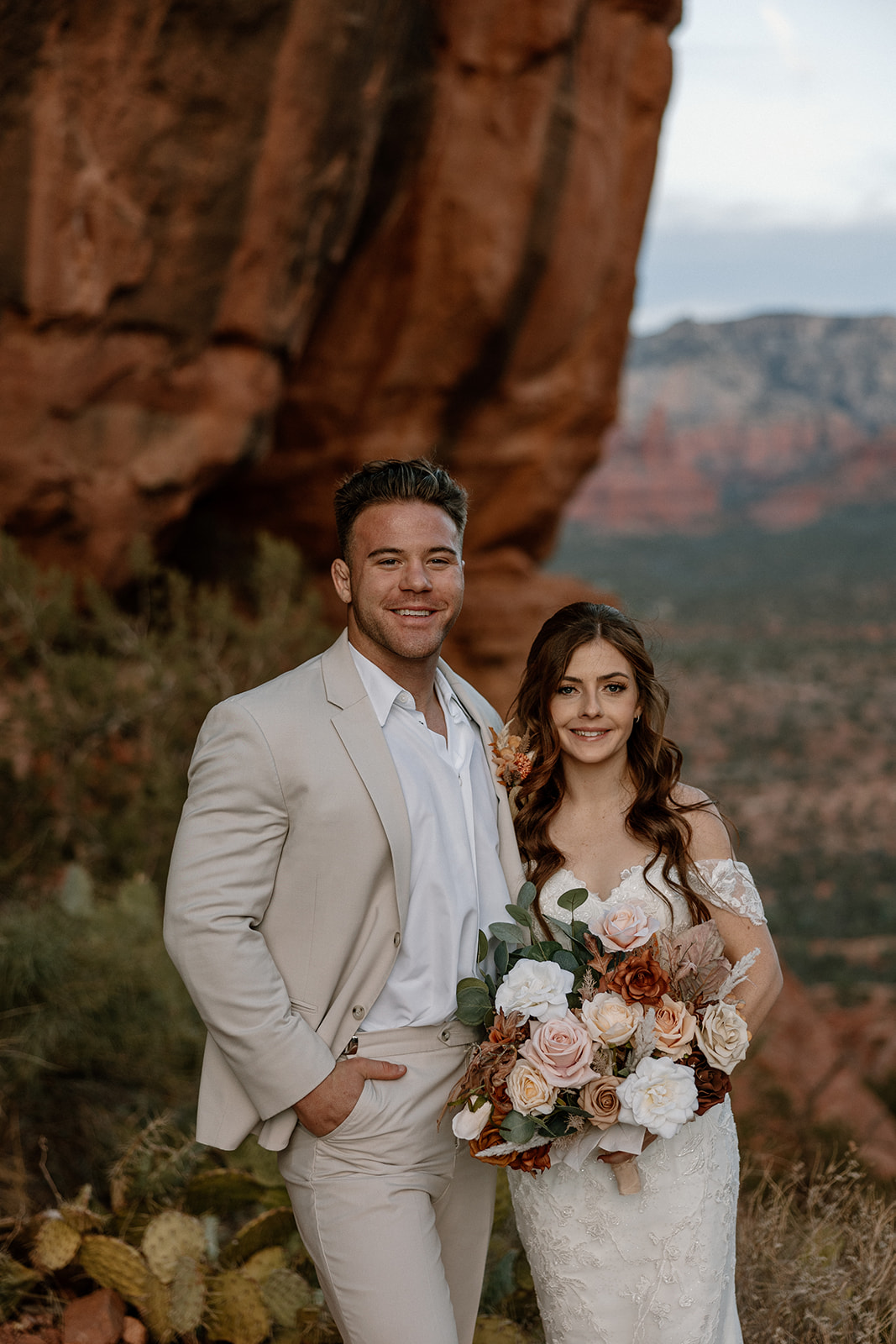 The newlyweds smile softly in front of towering canyon walls, holding a bouquet of dusty pink and ivory florals.