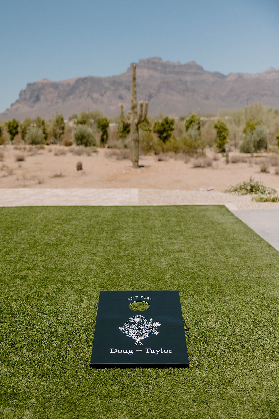 A personalized cornhole board sits on fresh green turf, with Superstition Mountain and towering saguaros creating a casual, epic reception backdrop.