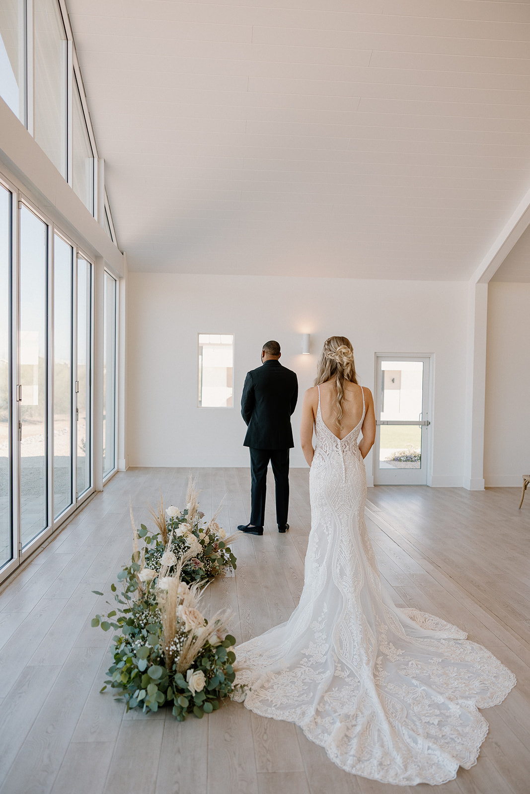 The bride walks toward her groom for their first look, her train trailing behind her, floral arrangements framing the soft wood floor.