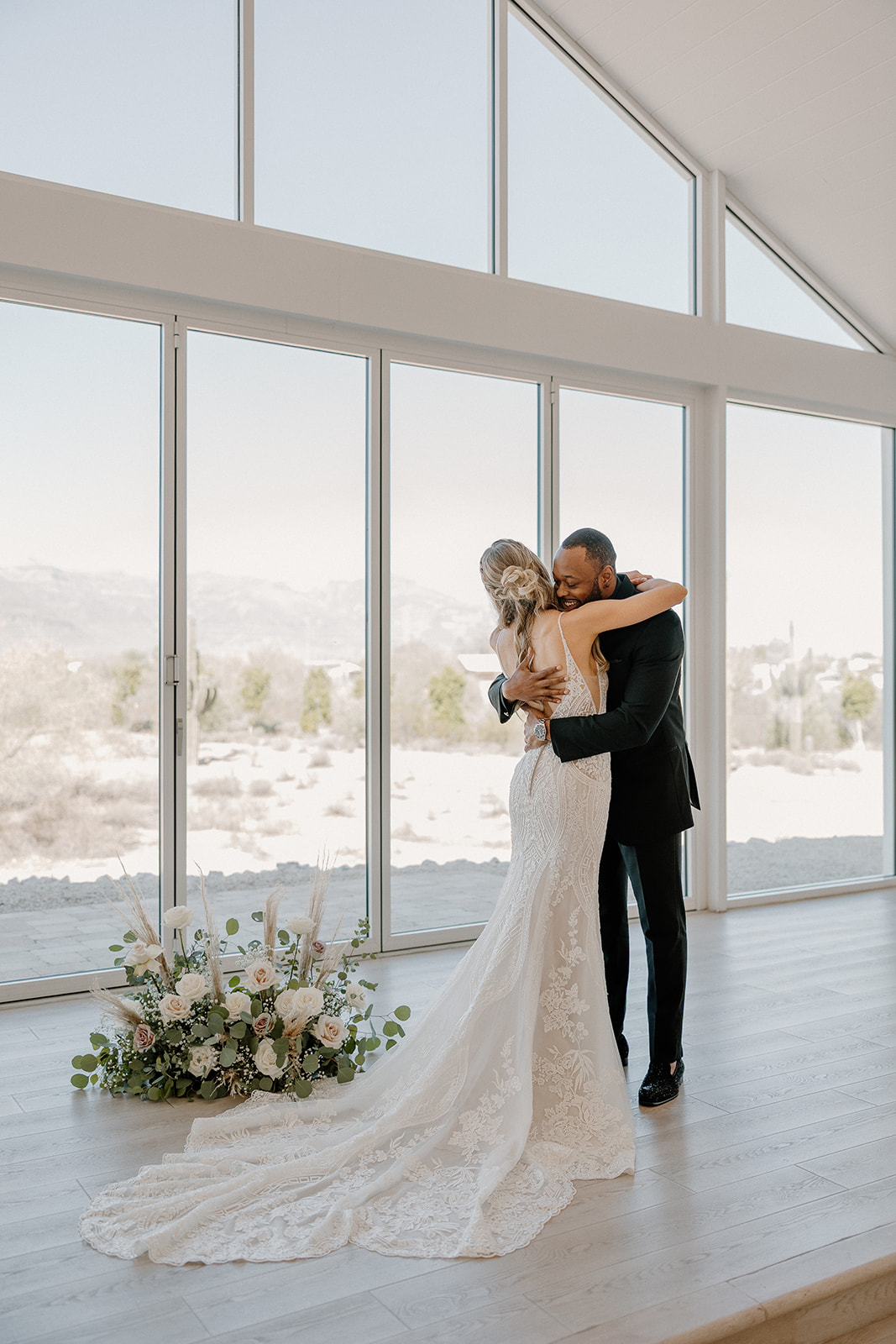 The couple shares a tight embrace during their first look, surrounded by floor-to-ceiling windows and soft desert light—desert view weddings romance in full force.