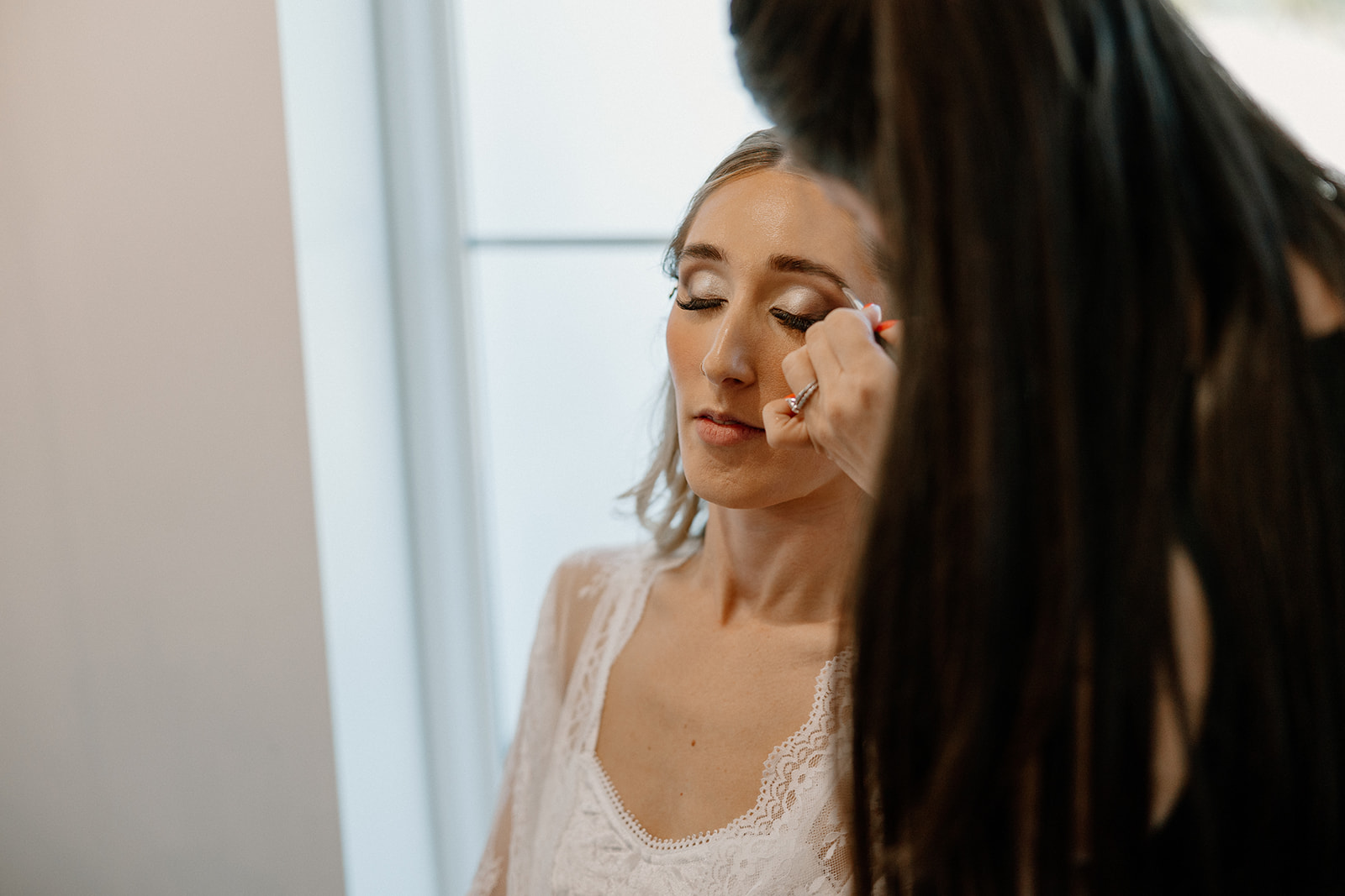 A makeup artist applies final touches to the bride’s eyes, her lace robe and peaceful expression capturing the calm before the ceremony.