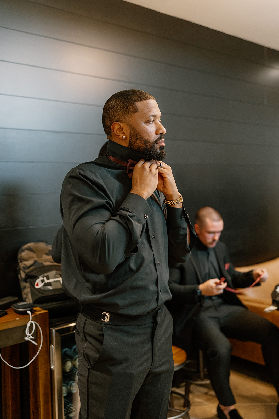 A groomsman adjusts his bowtie with laser focus while another relaxes in the background—final touches before the festivities begin.