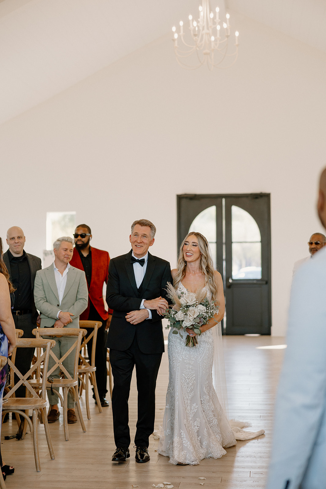 The bride beams while walking arm-in-arm with her father down the aisle, guests turning in their chairs to welcome her.