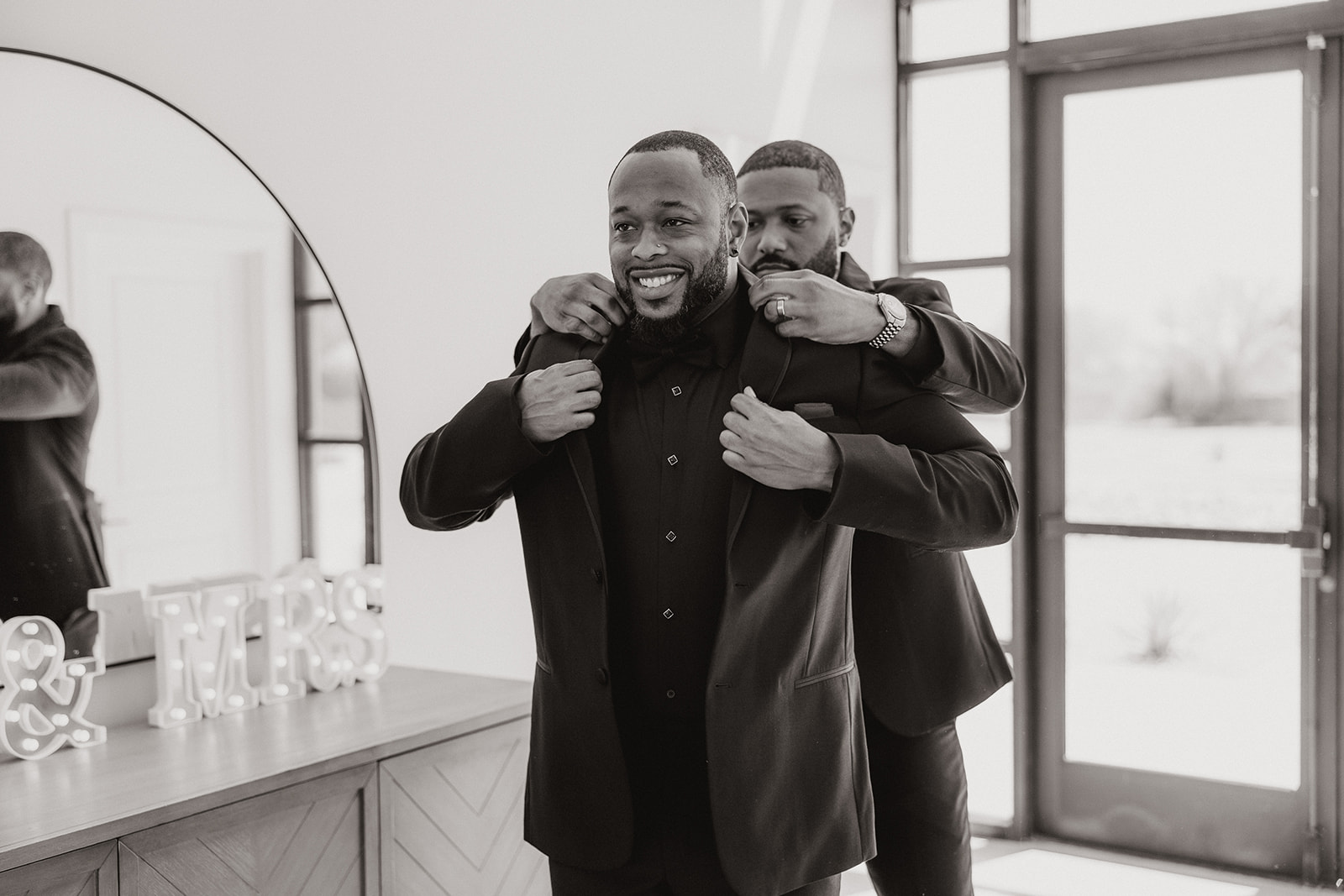A groomsman helps adjust the groom’s collar as he laughs in front of a mirror—black and white elegance with all the real energy.