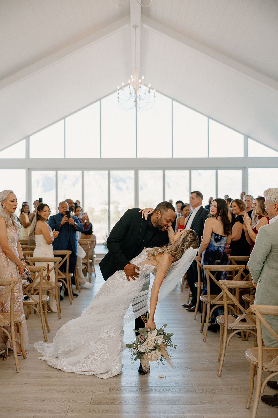 The groom dips the bride for a dramatic kiss at the end of the aisle as guests cheer them on—sunlight pouring into this glass-wrapped desert view weddings chapel.