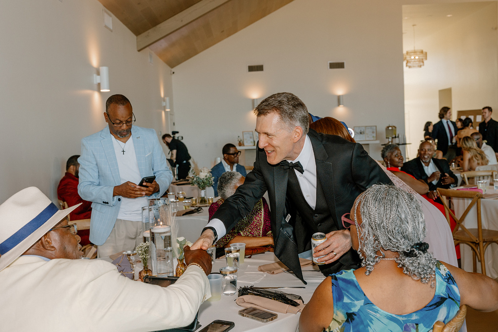 A sharply dressed guest in a tuxedo shares a warm handshake with an older gentleman in a white hat during cocktail hour—smiles all around.