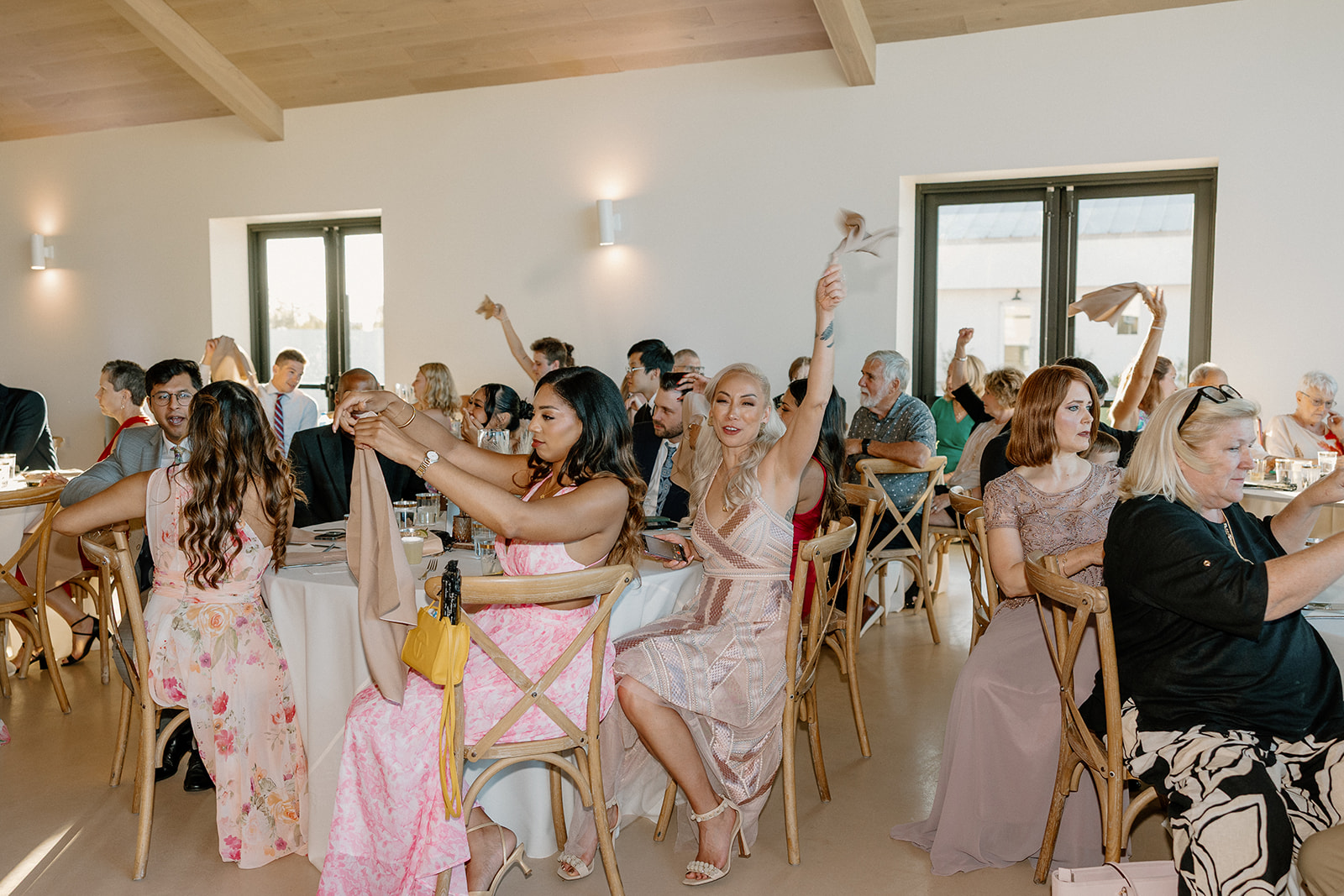 Reception guests wave napkins in the air mid-cheer, filling the modern indoor venue with celebratory energy and candid movement.