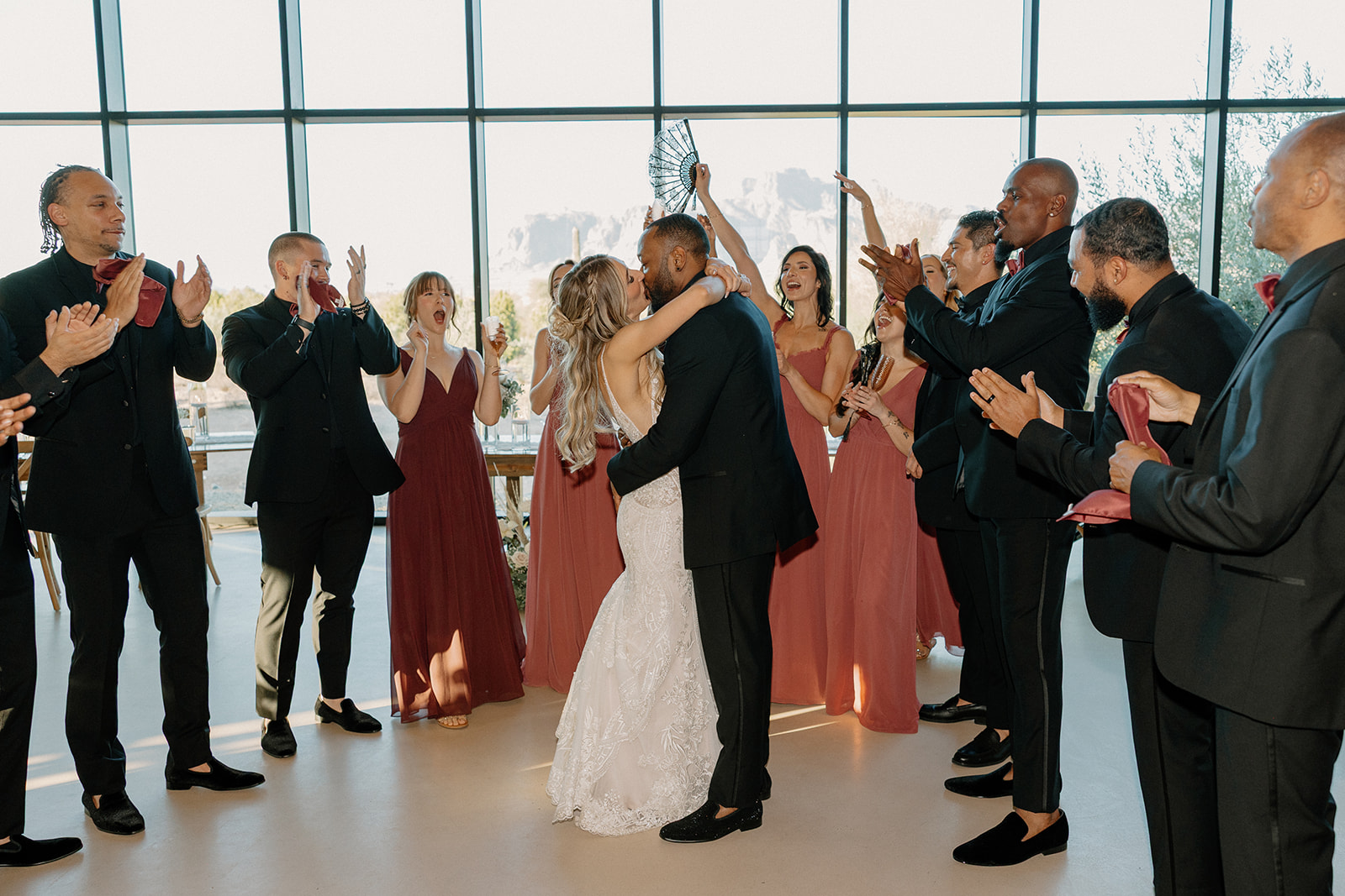 The newlyweds kiss while their wedding party surrounds them, cheering and clapping under the light-filled reception hall—an epic moment from their desert view weddings celebration.