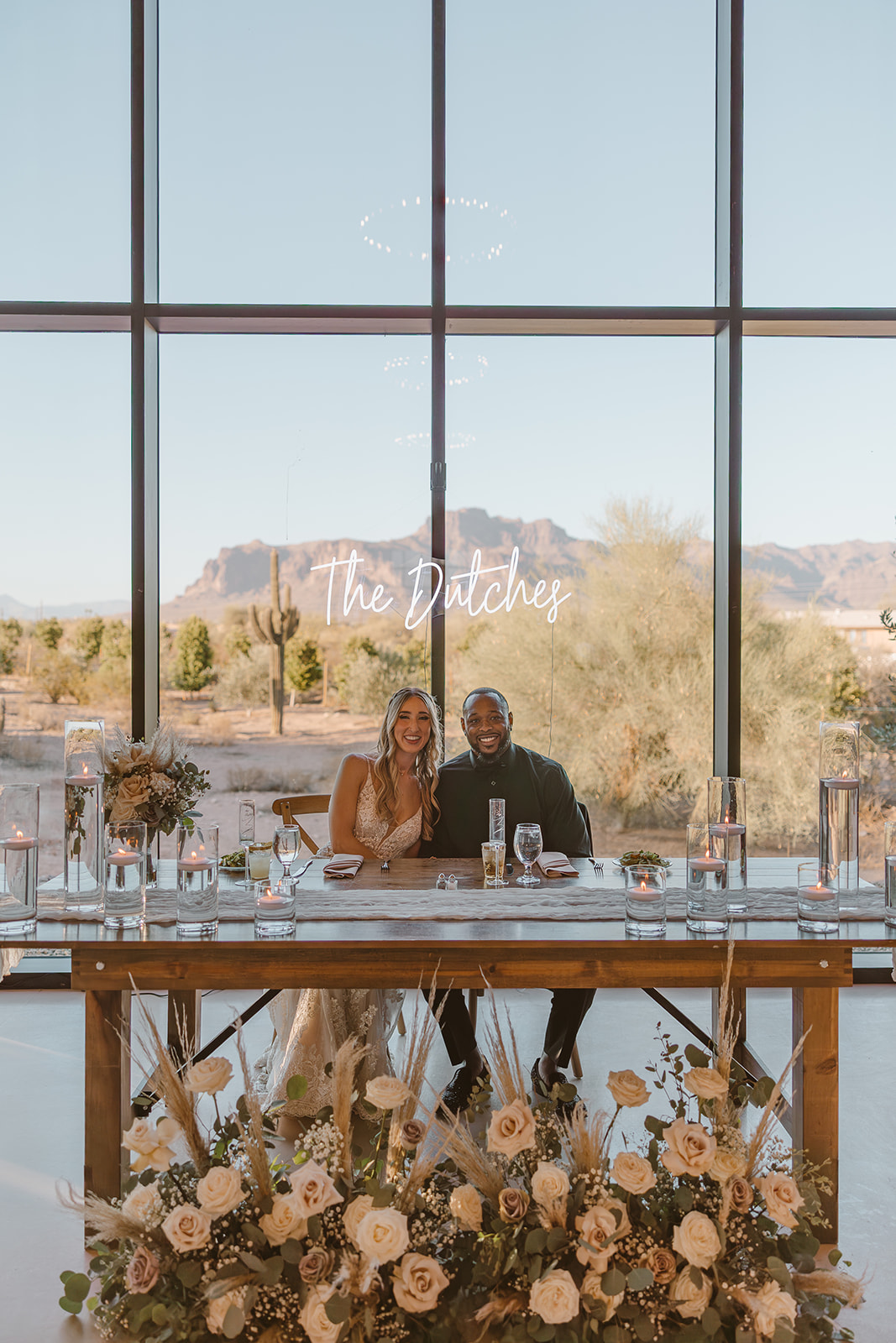 The bride and groom sit at their sweetheart table, framed by dreamy florals and desert peaks—“The Dutches” glowing behind them at this unforgettable desert view weddings reception.