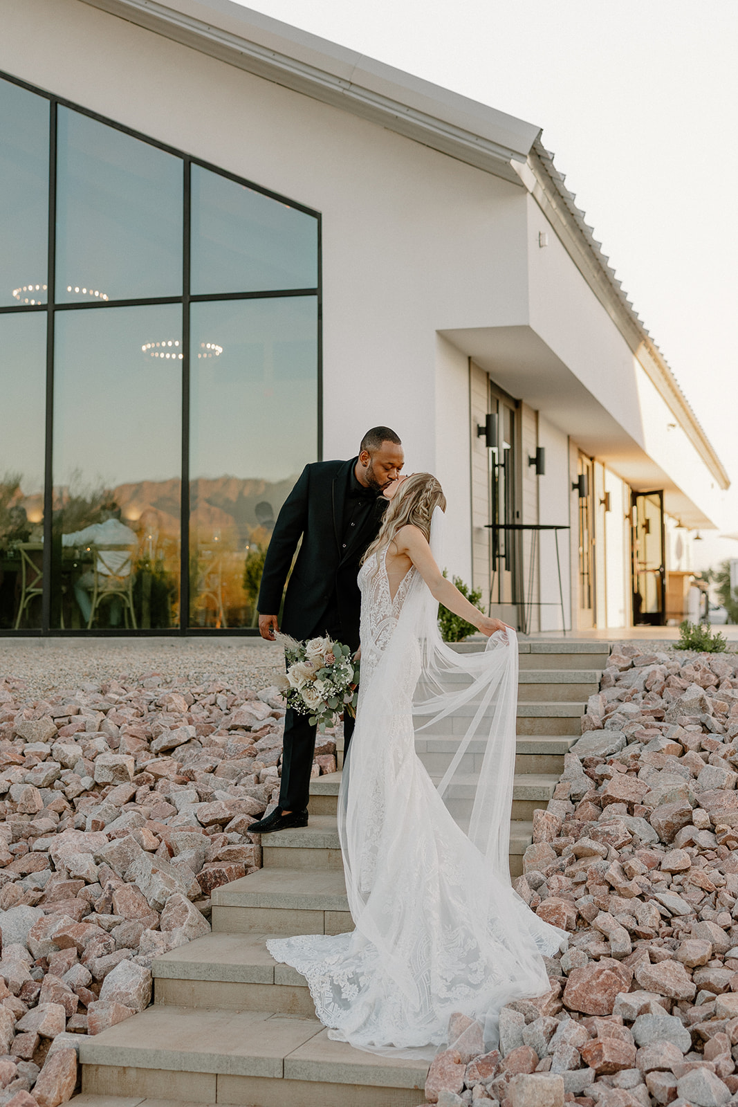 At the top of the venue stairs, the groom sweetly kisses the bride’s hand as she gathers her dress—quiet romance, Arizona style.