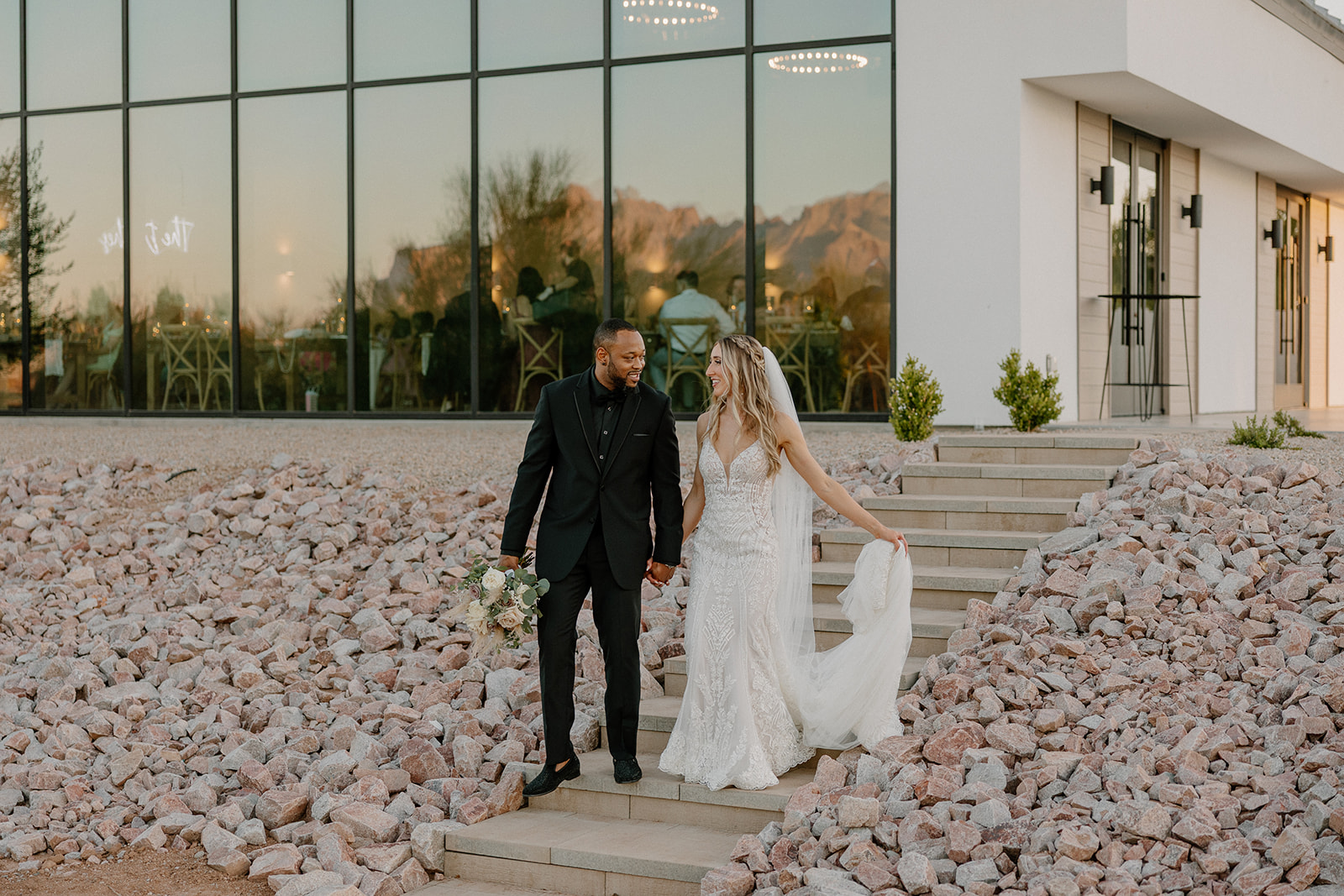 The bride and groom walk hand-in-hand down stone steps outside the venue, glowing in golden hour light with reflections of desert peaks behind them—peak desert view weddings energy.