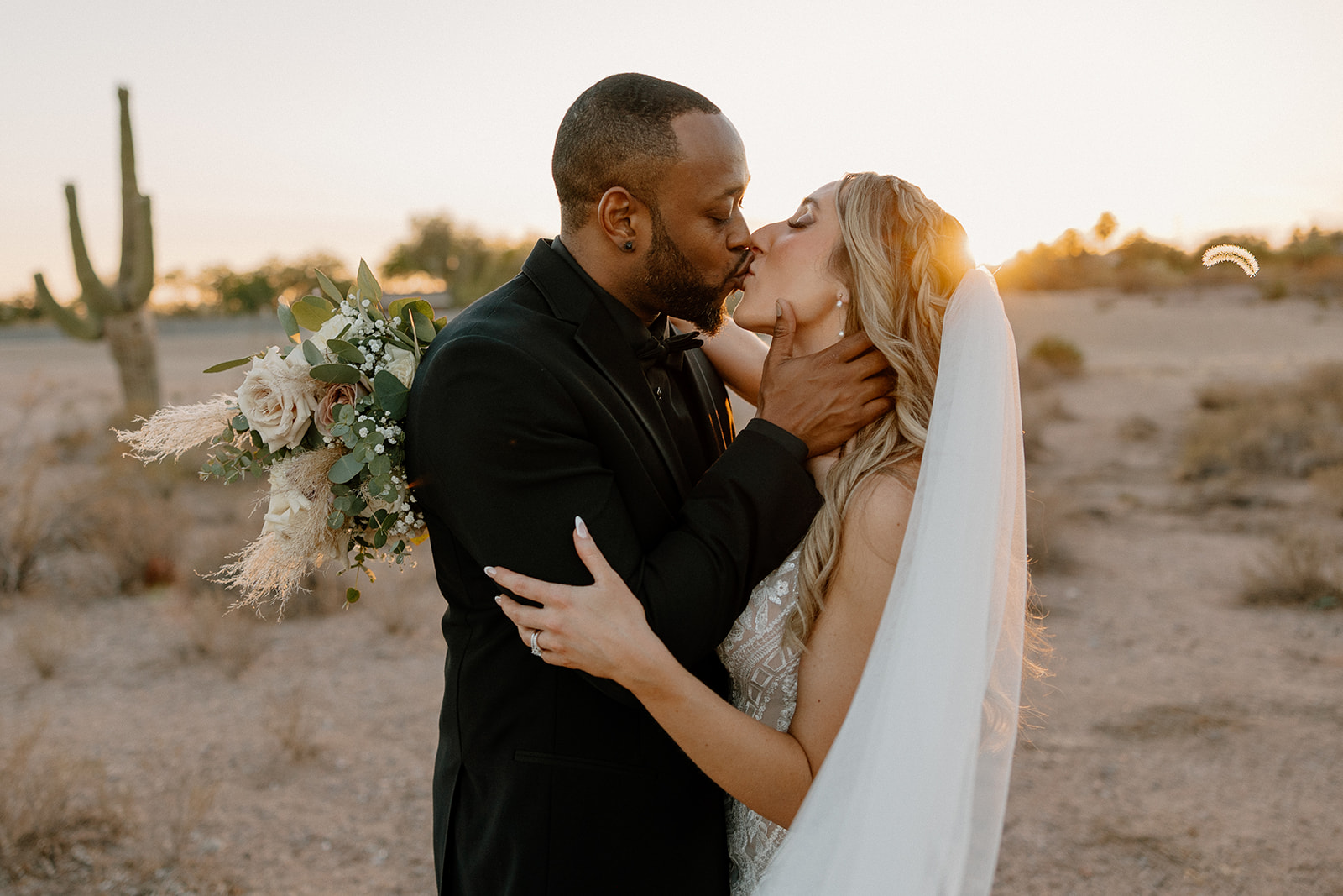 Golden light spills across the desert as the couple shares a romantic kiss—cactus silhouettes and soft shadows setting the scene.
