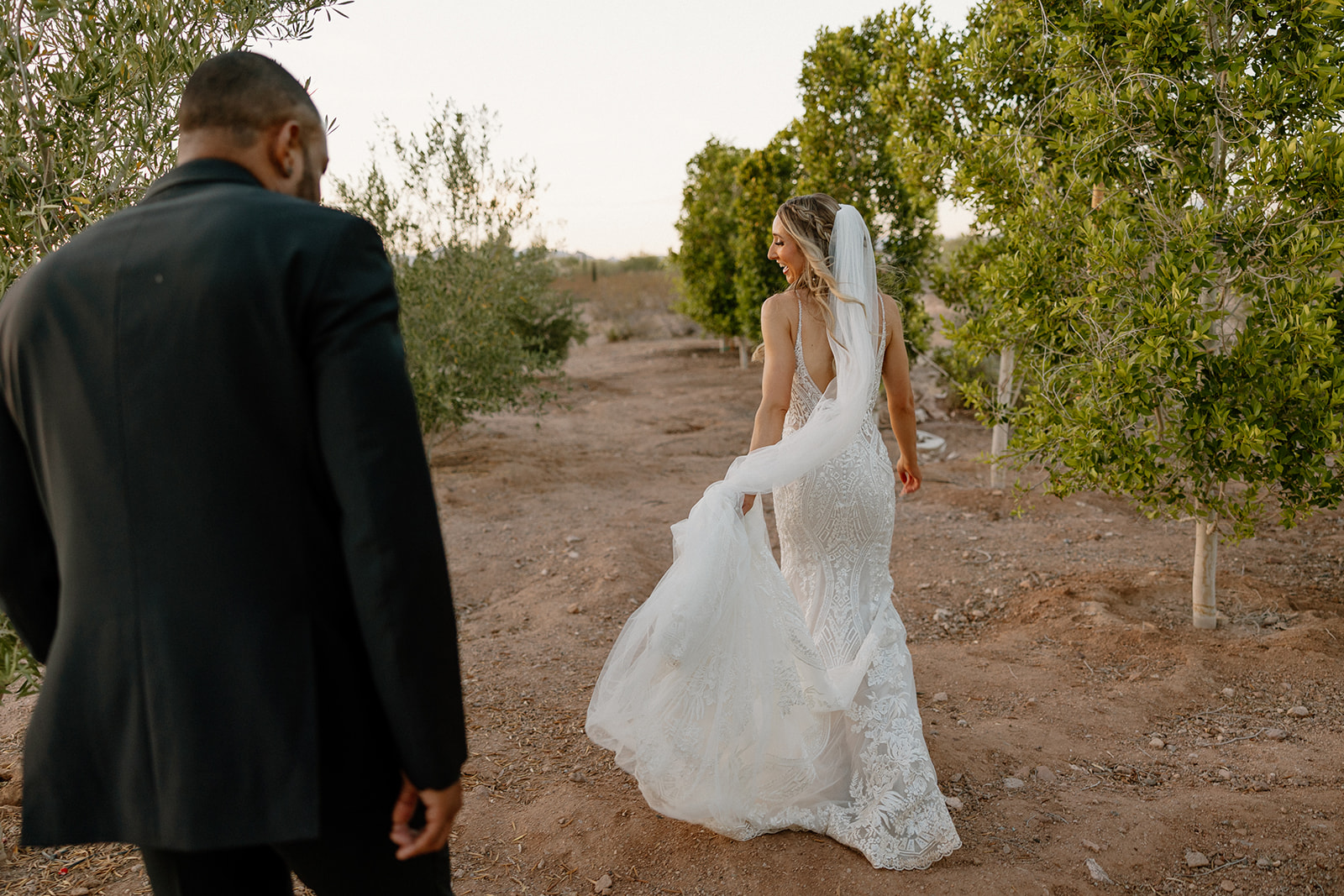 The bride glances back with a soft smile as she leads the groom through an orchard path, her lace train trailing through the Arizona dirt.