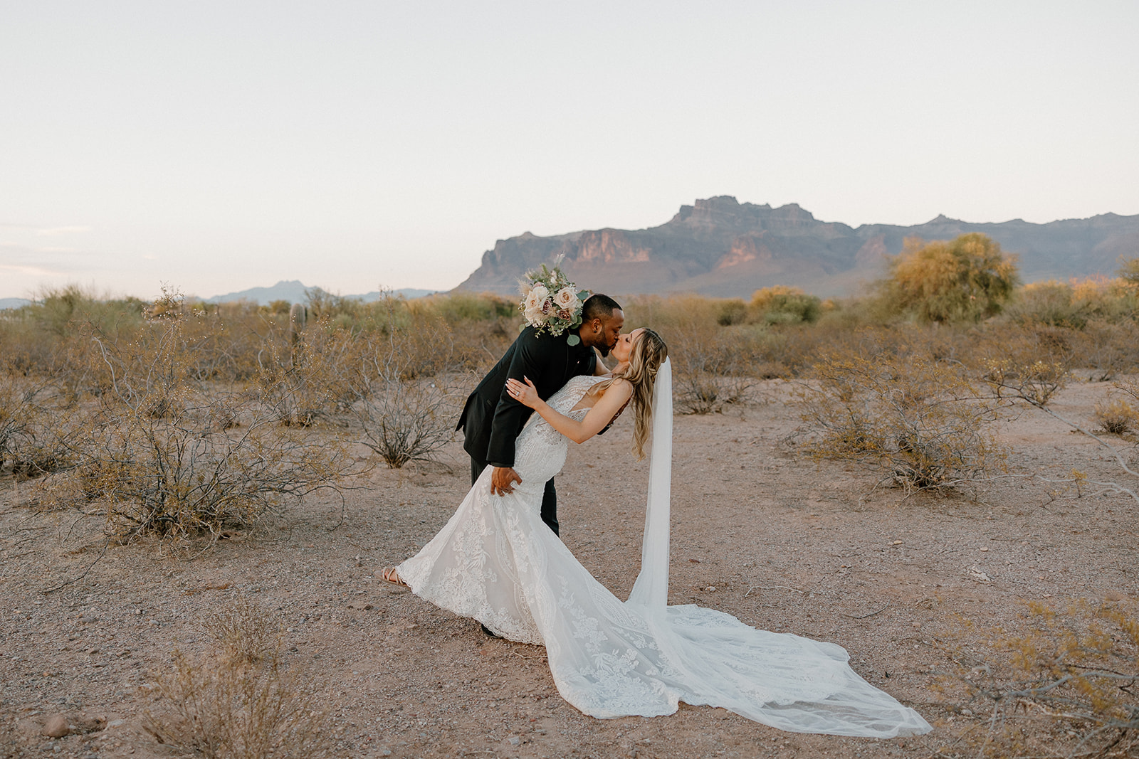 The groom dips the bride for a kiss in front of the Superstition Mountains—lace gown flowing, florals in hand, and full desert view weddings magic in motion.
