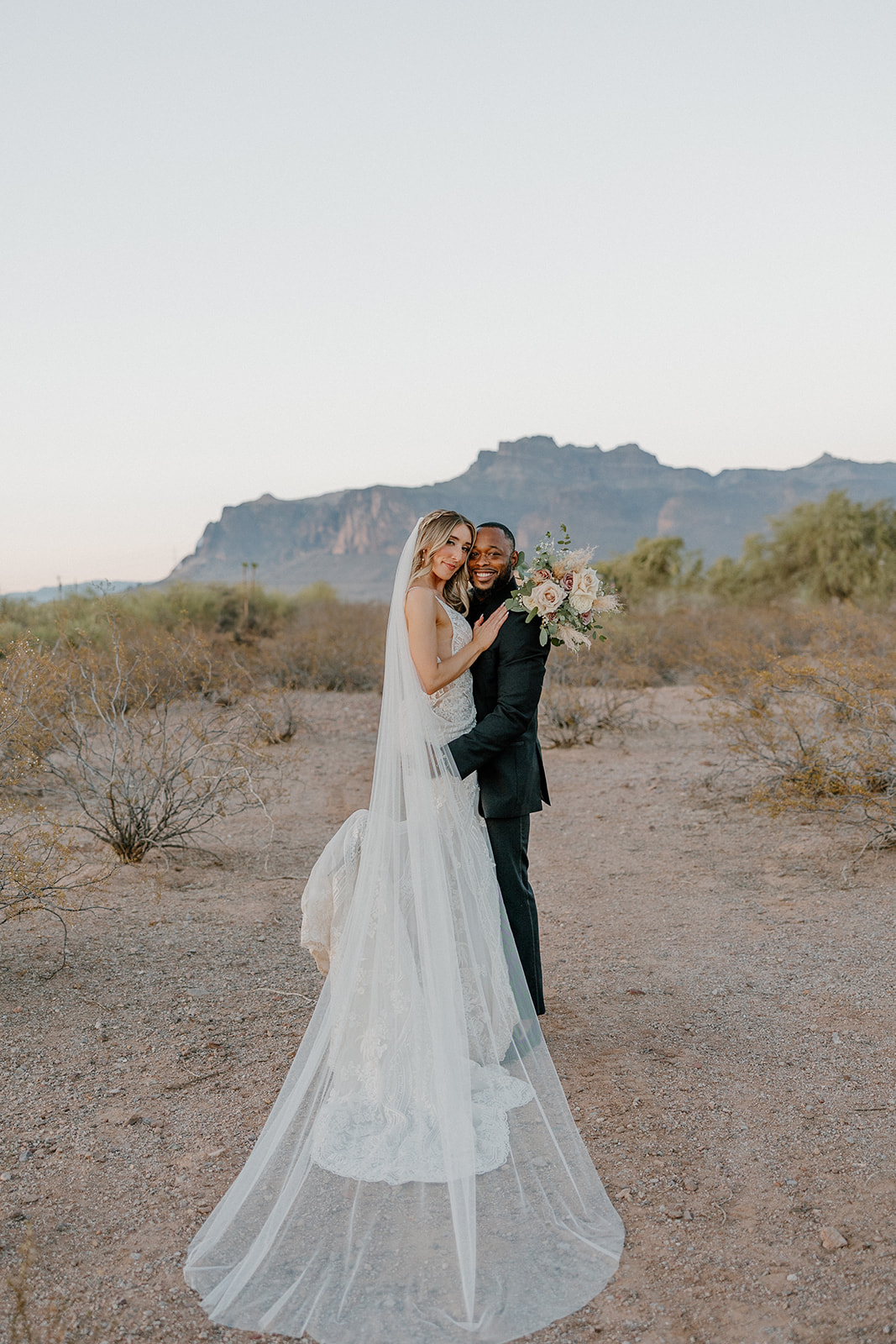 The couple cuddles up in a warm embrace as the bride’s veil floats behind them, desert mountains rising softly in the distance.