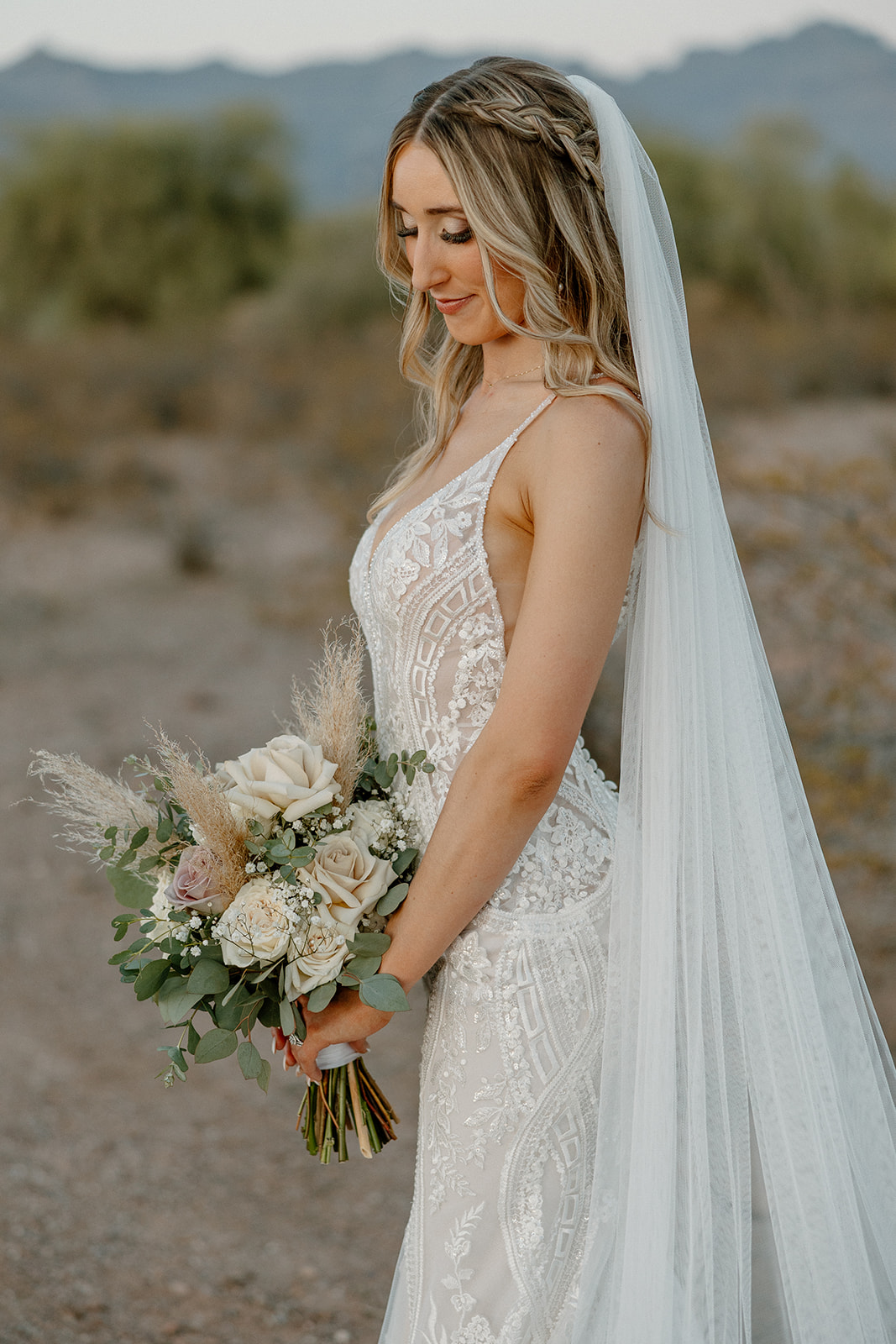 The bride stands in profile, gazing at her pampas-filled bouquet, gown catching golden hour light on this desert view weddings day.