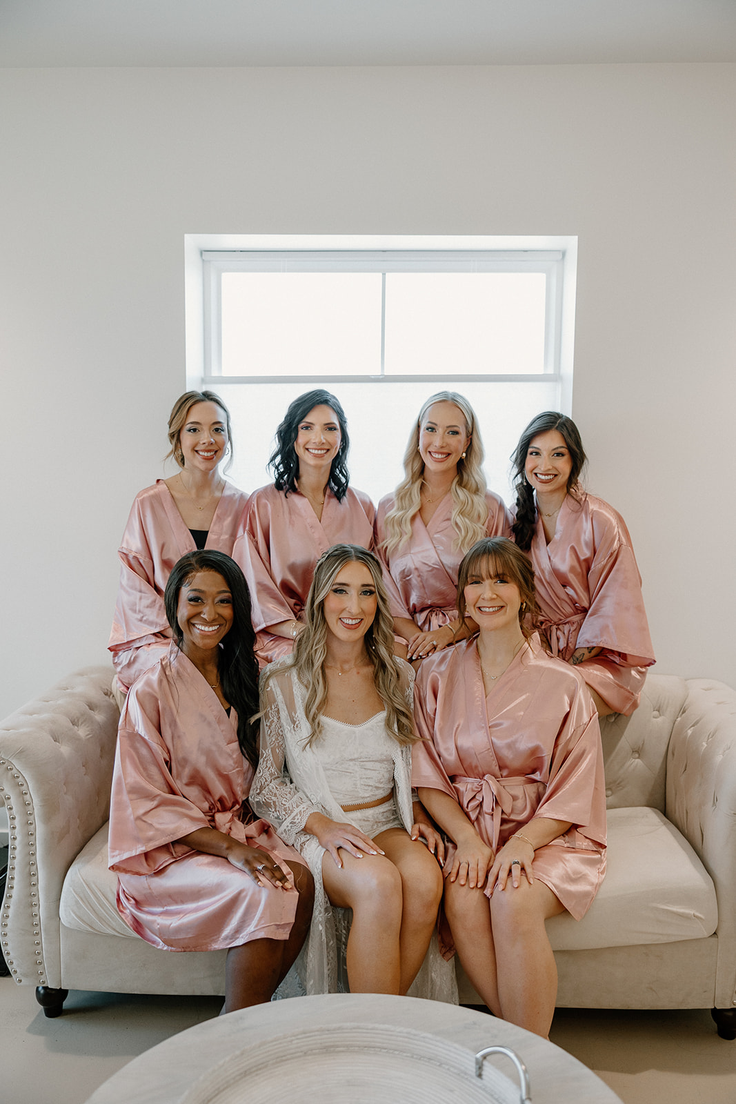 The bride and her bridesmaids sit in matching pink satin robes, smiling wide on a cozy couch before the day officially begins.