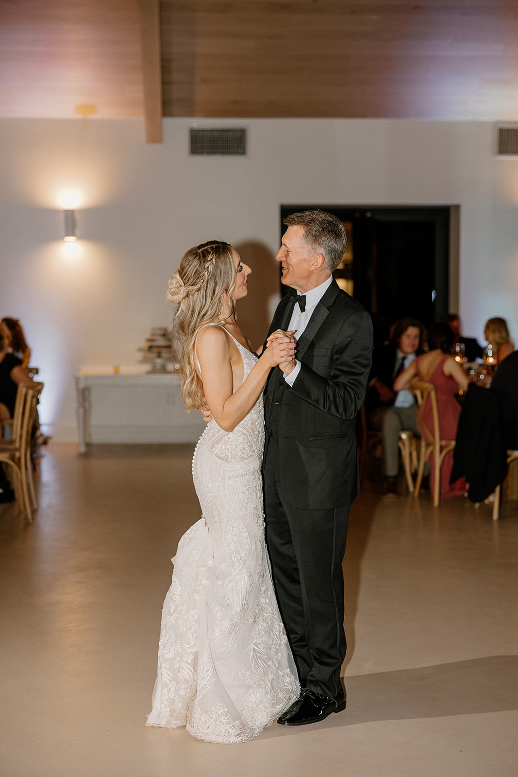 The bride holds hands with her father during a sentimental dance, her lace gown glowing in the soft evening light.