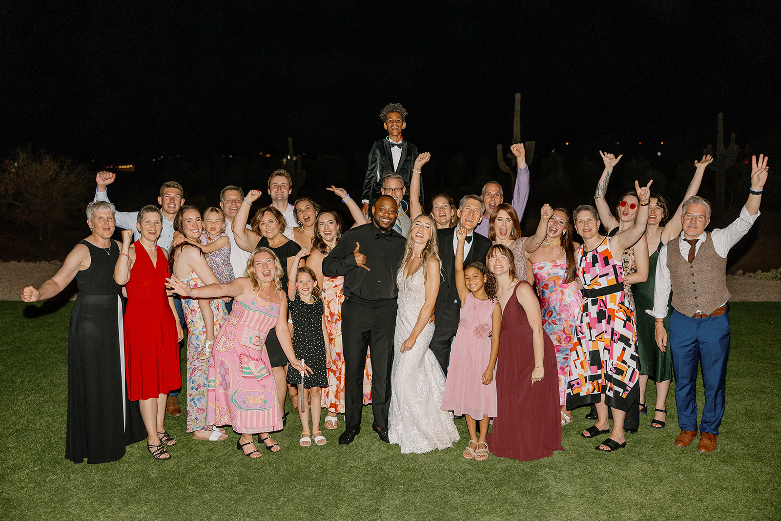 The bride and groom stand surrounded by an excited crowd of friends and family, arms raised in celebration against the cactus-lined night sky—pure joy at this desert view weddings celebration.
