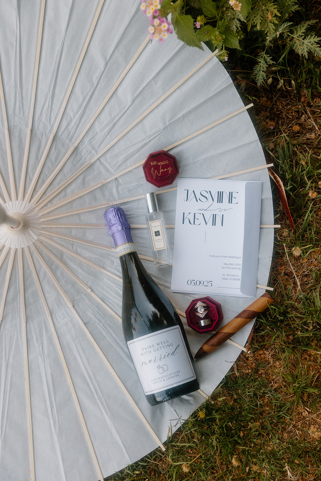 A Western wedding flatlay set atop a white paper parasol, including champagne, two red velvet ring boxes, perfume, a wedding program for Jasmine and Kevin, and a rolled cigar, styled with thoughtful rustic details.