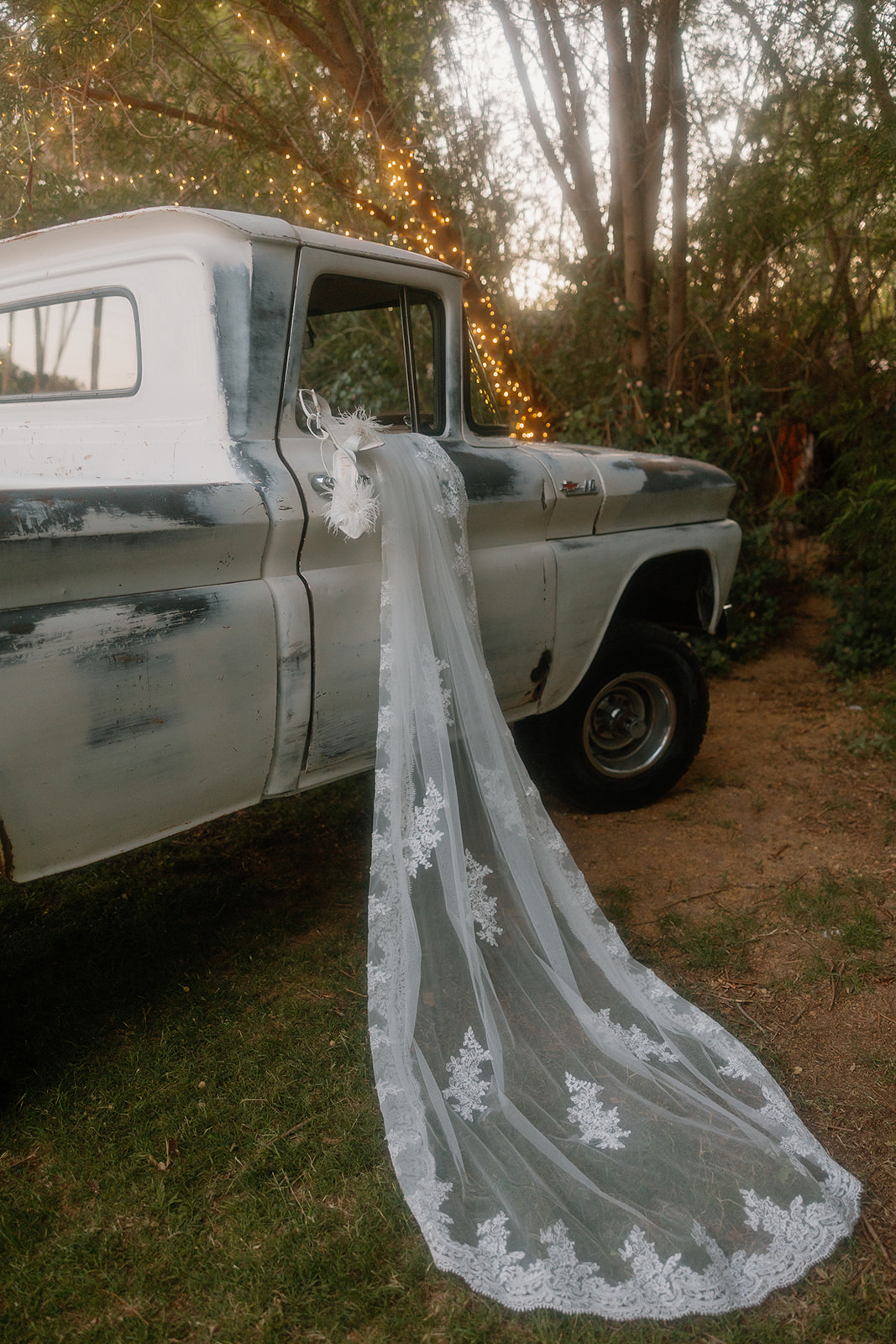 The bride’s veil and feathered heels hang delicately from the door of a vintage white and black pickup, the veil trailing dramatically down to the grass—Western wedding textures at their finest.