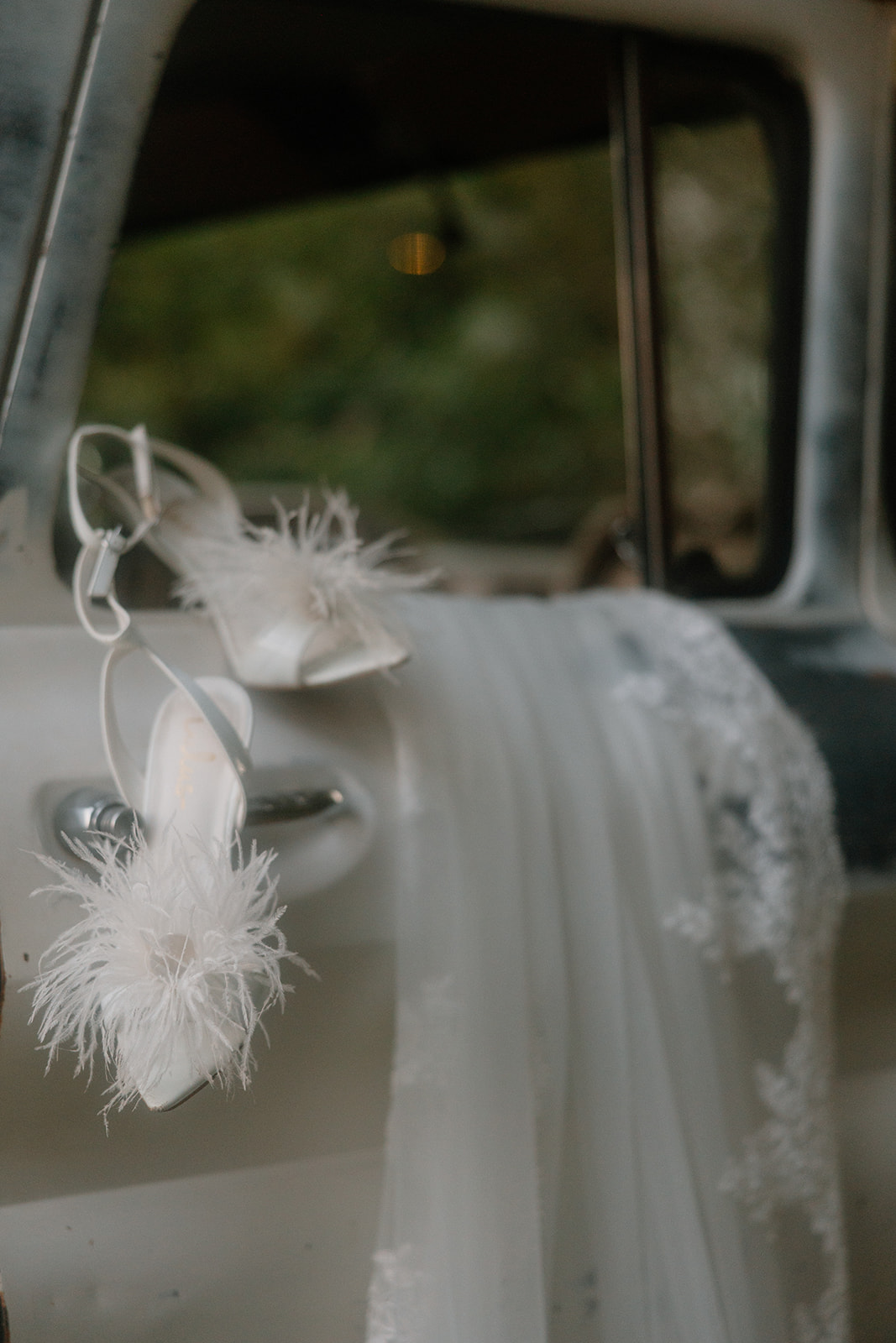 Feathered white bridal heels hang from the handle of a vintage truck door, next to delicate lace from a wedding veil—effortlessly mixing Western grit with modern bridal glam.