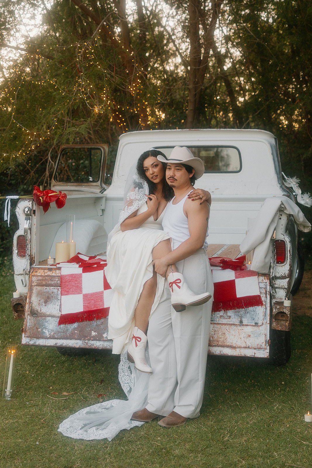 A bold Western wedding portrait with the bride lifted into the groom’s arms, rocking white boots with red ribbon details and her veil cascading behind. A vintage truck and candlelight complete the scene.