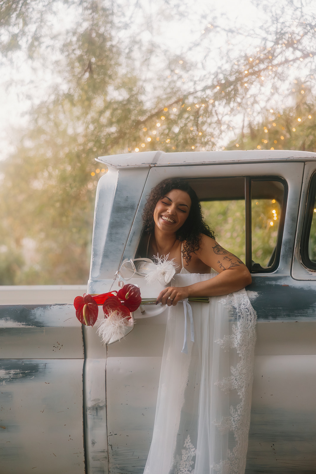 The bride beams with joy, leaning out the window of a vintage truck holding a bouquet of red anthuriums and white feathers, her lace veil catching soft morning light.