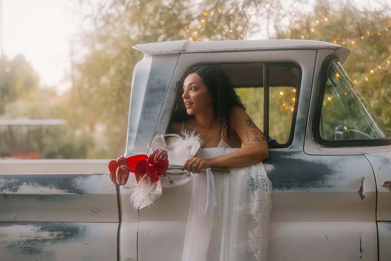 A bride leans dreamily out of a weathered white-and-blue vintage pickup truck window, holding a bold bouquet of red anthuriums and white feathers, surrounded by soft golden-hour light and string lights in the trees.