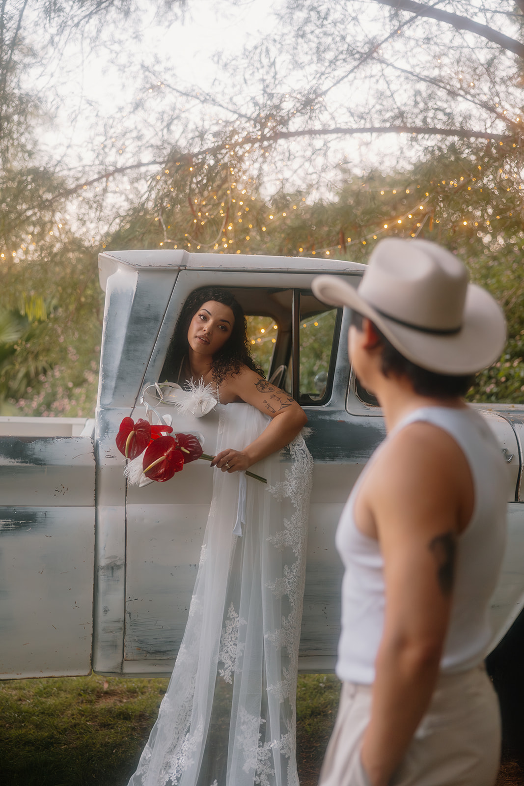 A bride with a dreamy expression leans out of a weathered vintage truck holding a red bouquet, while her groom in a cowboy hat gazes at her with admiration during their rustic Western wedding.