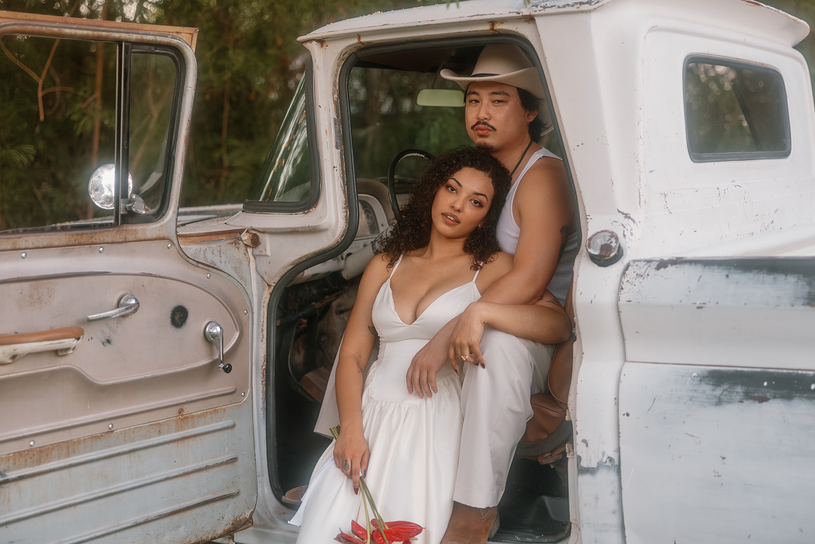 Bride and groom sitting in the cab of a vintage white pickup truck during their Western wedding, with the bride leaning softly against the groom’s chest holding a bold red bouquet, and the groom in a cowboy hat gazing ahead. The open truck door and worn textures add rustic charm to the intimate moment.