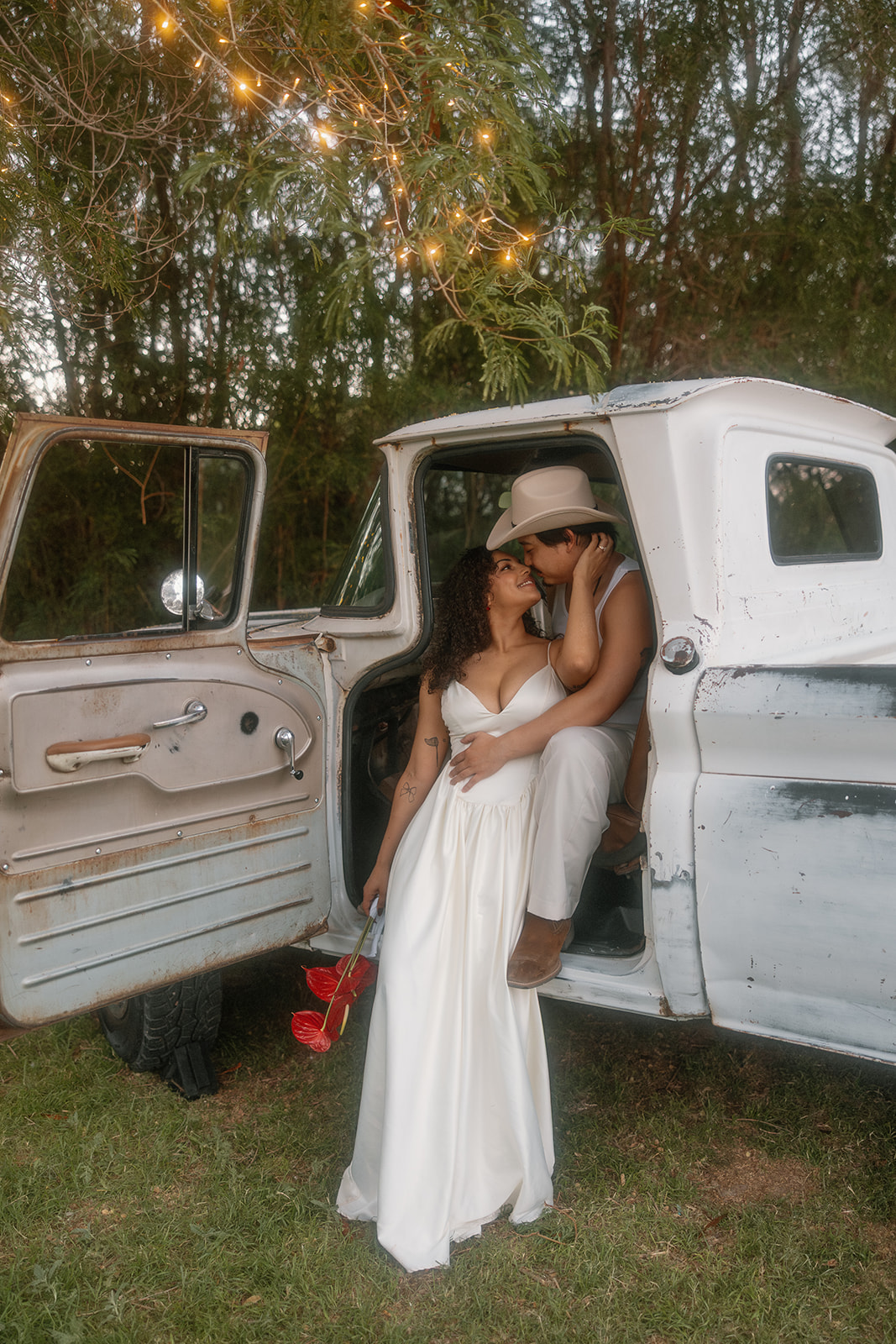 A Western bride and groom embrace and kiss at the open door of a vintage truck, surrounded by trees and string lights. Her dress and bouquet of red anthuriums contrast beautifully with his cowboy boots and hat.