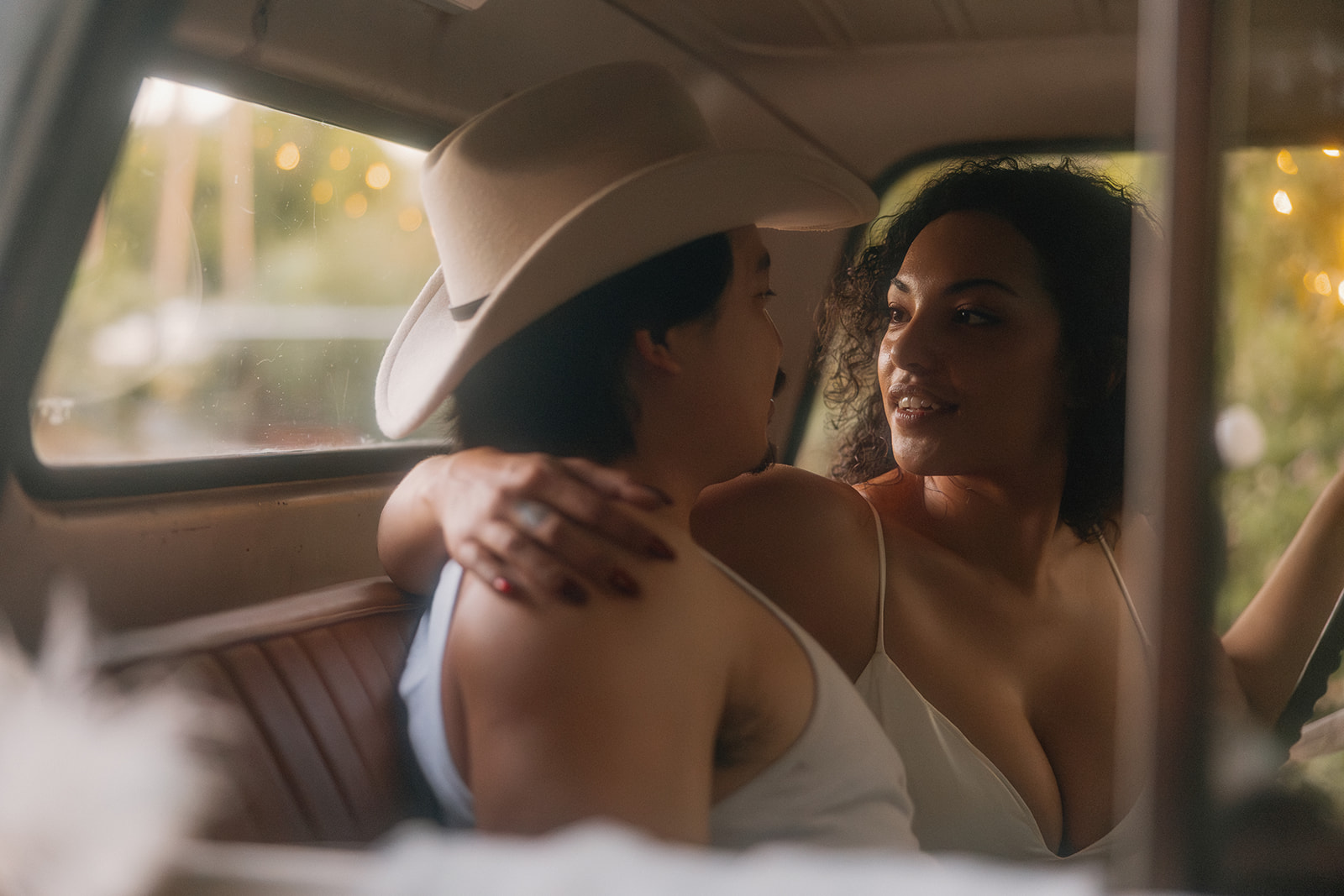 A romantic close-up of a newlywed couple inside a vintage truck, the bride gazing into her partner’s eyes while he wears a cowboy hat and tank top. The moment is intimate, glowing with soft light and rustic charm.