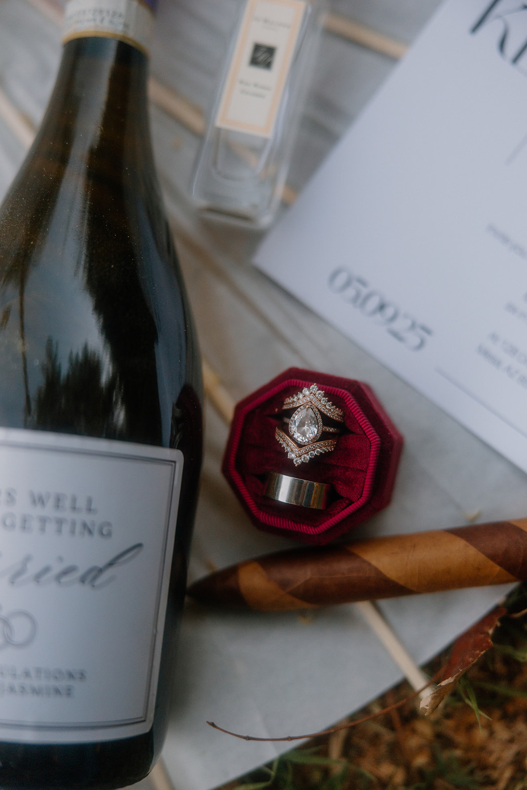 A styled flatlay featuring a bottle of champagne, wedding rings in a velvet box, a rolled cigar, a Jo Malone perfume bottle, and a minimalist wedding invite—capturing the luxe-meets-Western wedding aesthetic.