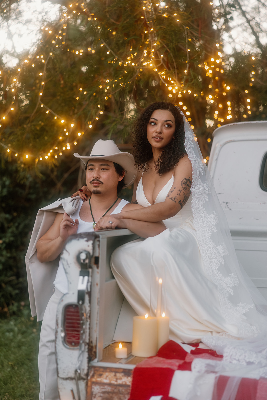 A bride lounges on the tailgate of a vintage pickup while her groom stands protectively beside her, wrapped in her veil. Candles, a checkered blanket, and twinkle lights bring a romantic Western wedding vibe.