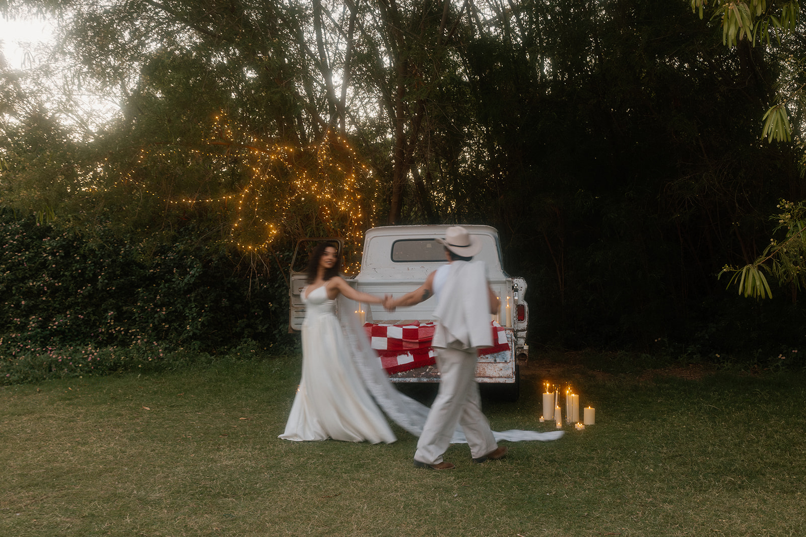 A motion-blurred image of a couple twirling hand-in-hand in front of a vintage truck, captured during their Western wedding at sunset, surrounded by candles and glowing tree lights.