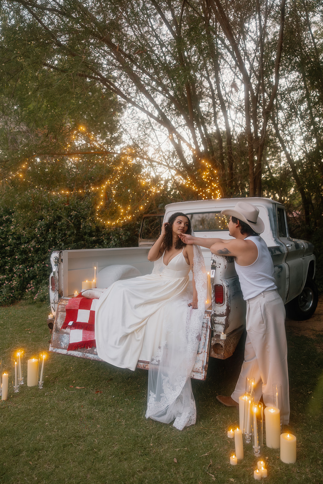 The bride sits in the bed of a vintage truck in her flowy dress and veil, gently touching her groom's face as he stands close. Twinkle lights and candles fill the intimate Western wedding scene.