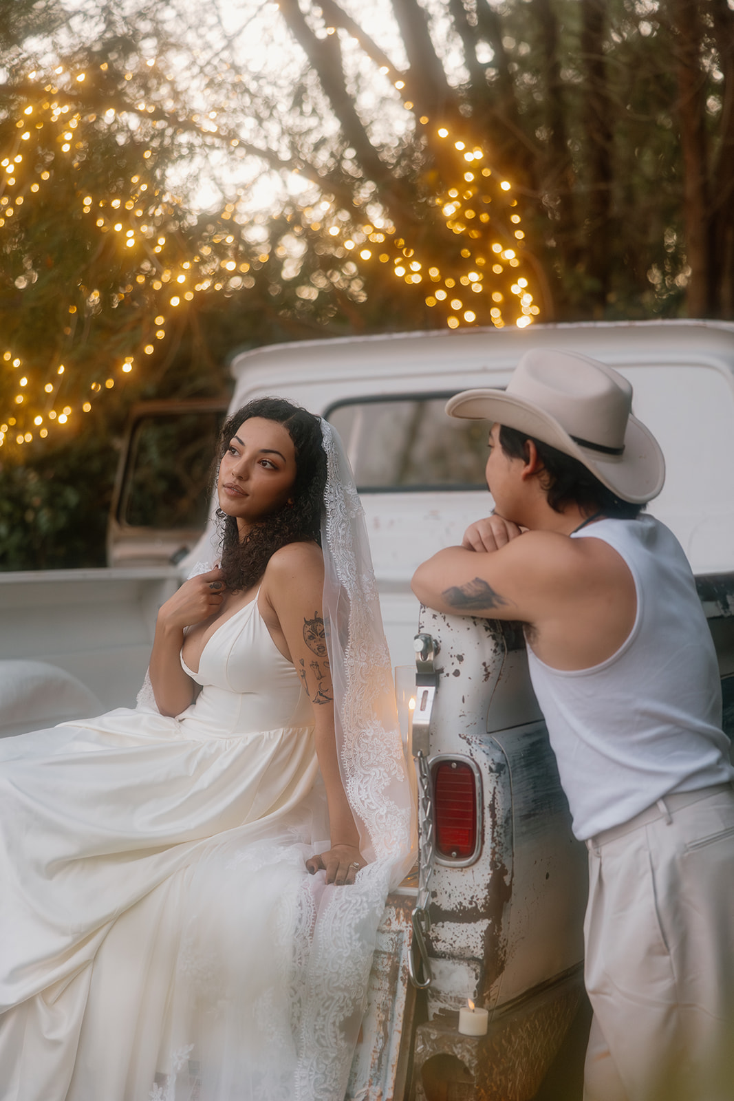 Golden hour glow bathes the bride as she sits on the truck bed in her wedding dress and veil, looking dreamy while her groom leans on the side of the truck, both framed by fairy lights.