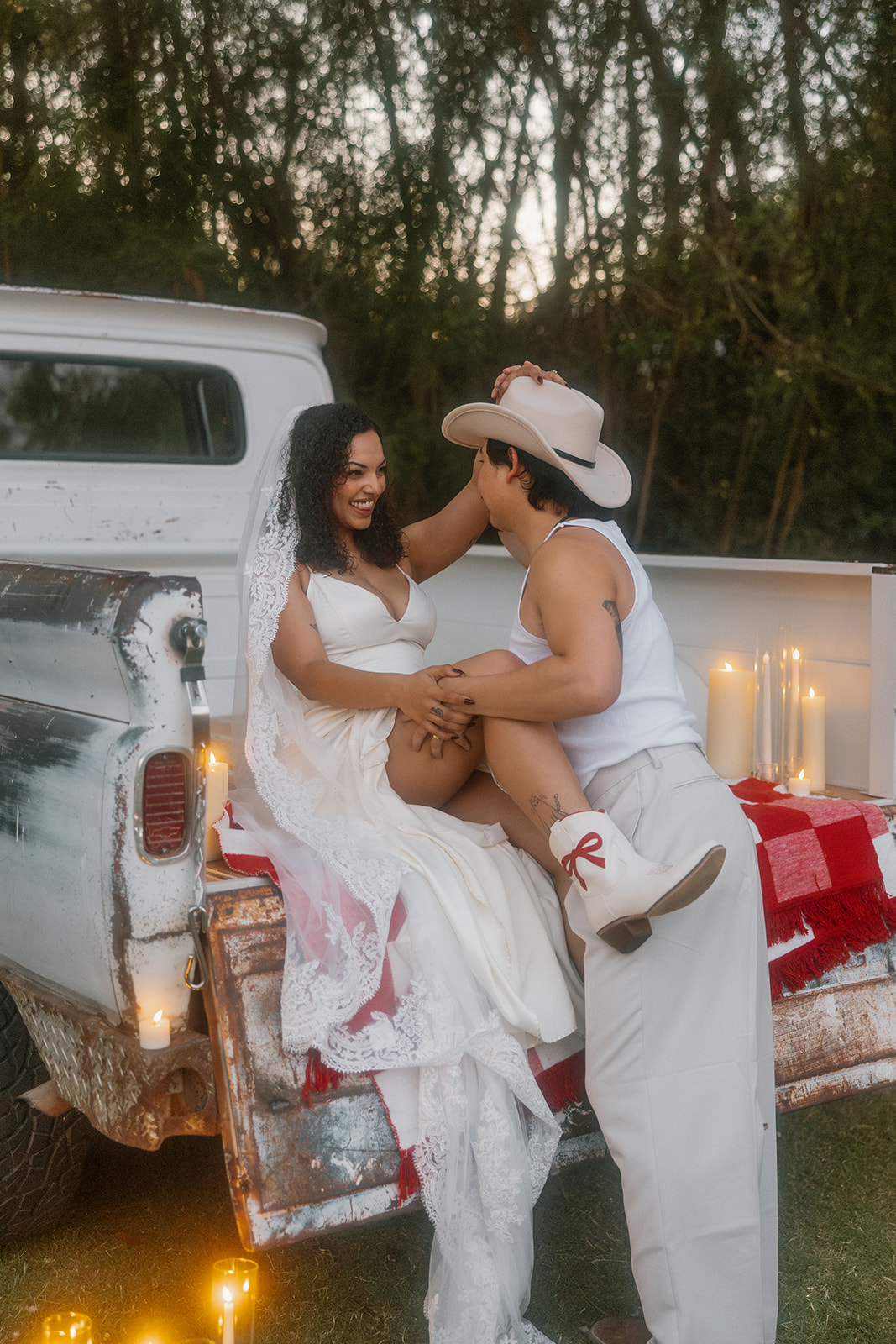 The bride sits on the edge of a vintage pickup truck bed, her leg lifted playfully on the groom’s lap as they share a flirtatious moment, surrounded by candles and twinkling lights.