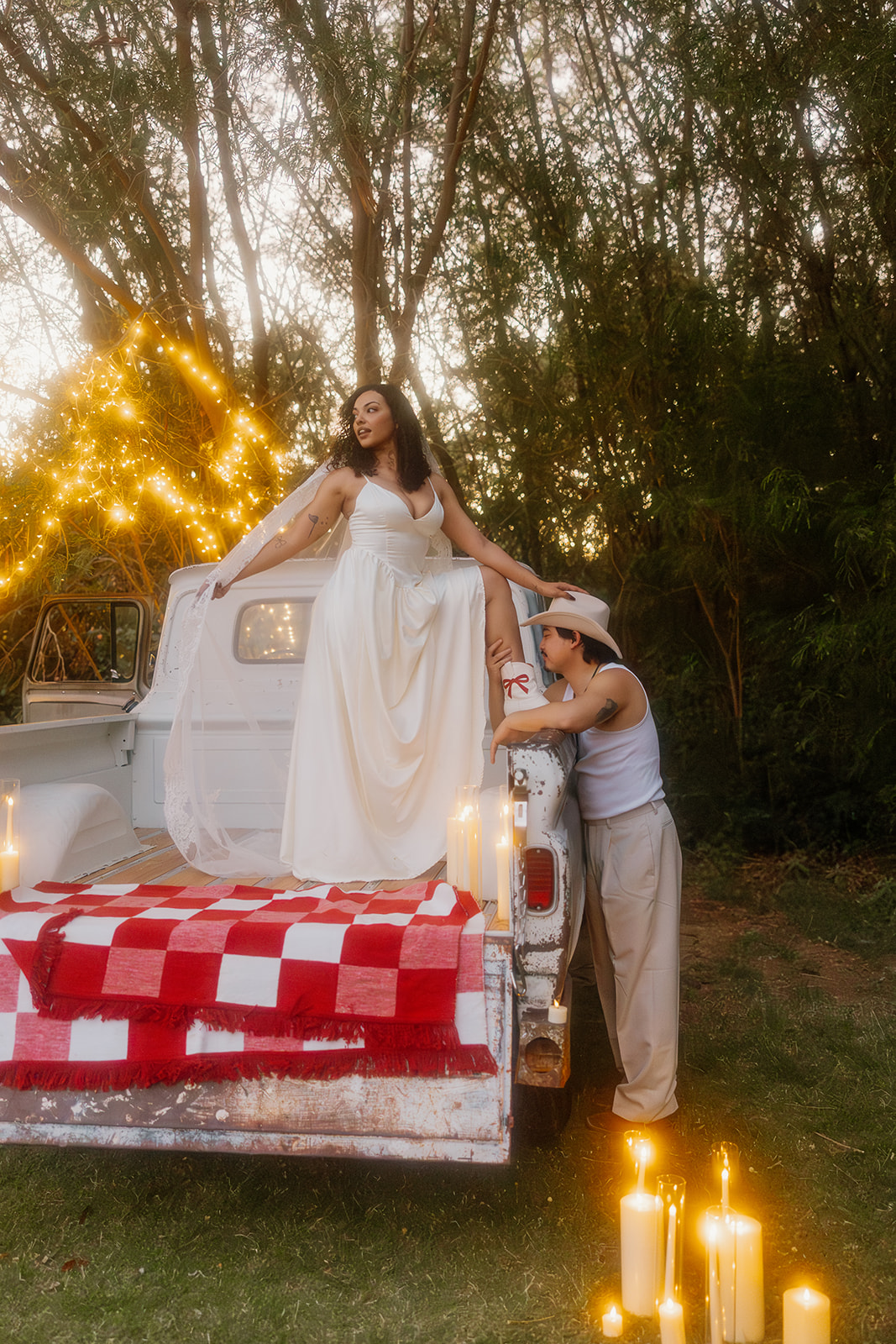 A romantic Western wedding moment with the bride standing barefoot in the truck bed, holding her groom’s hand as he gazes up at her, surrounded by candlelight and glowing string lights in the trees.