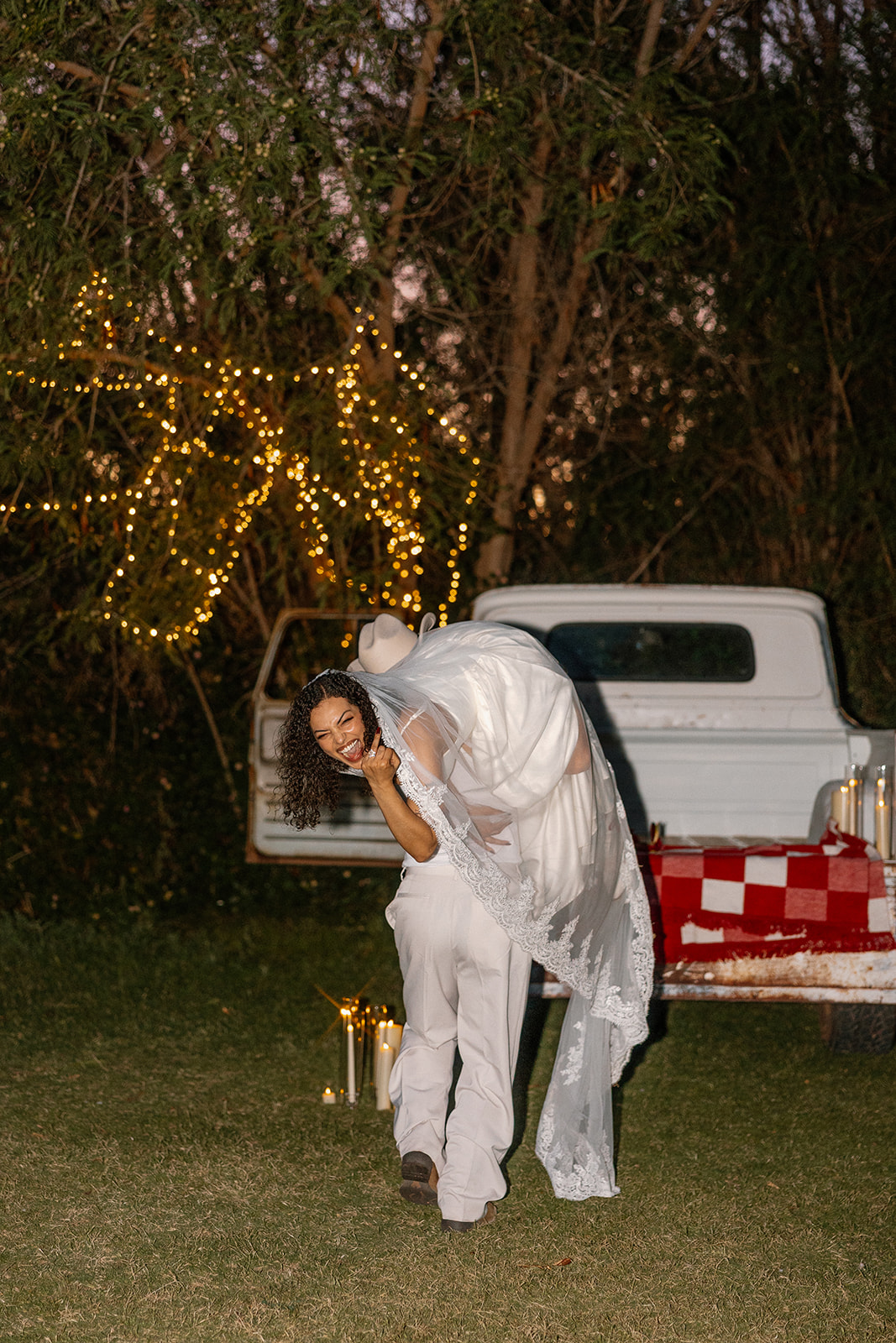 A playful Western wedding moment with the groom carrying the bride over his shoulder in front of a vintage truck. Candles and string lights add ambiance to this fun-loving, woodsy scene.