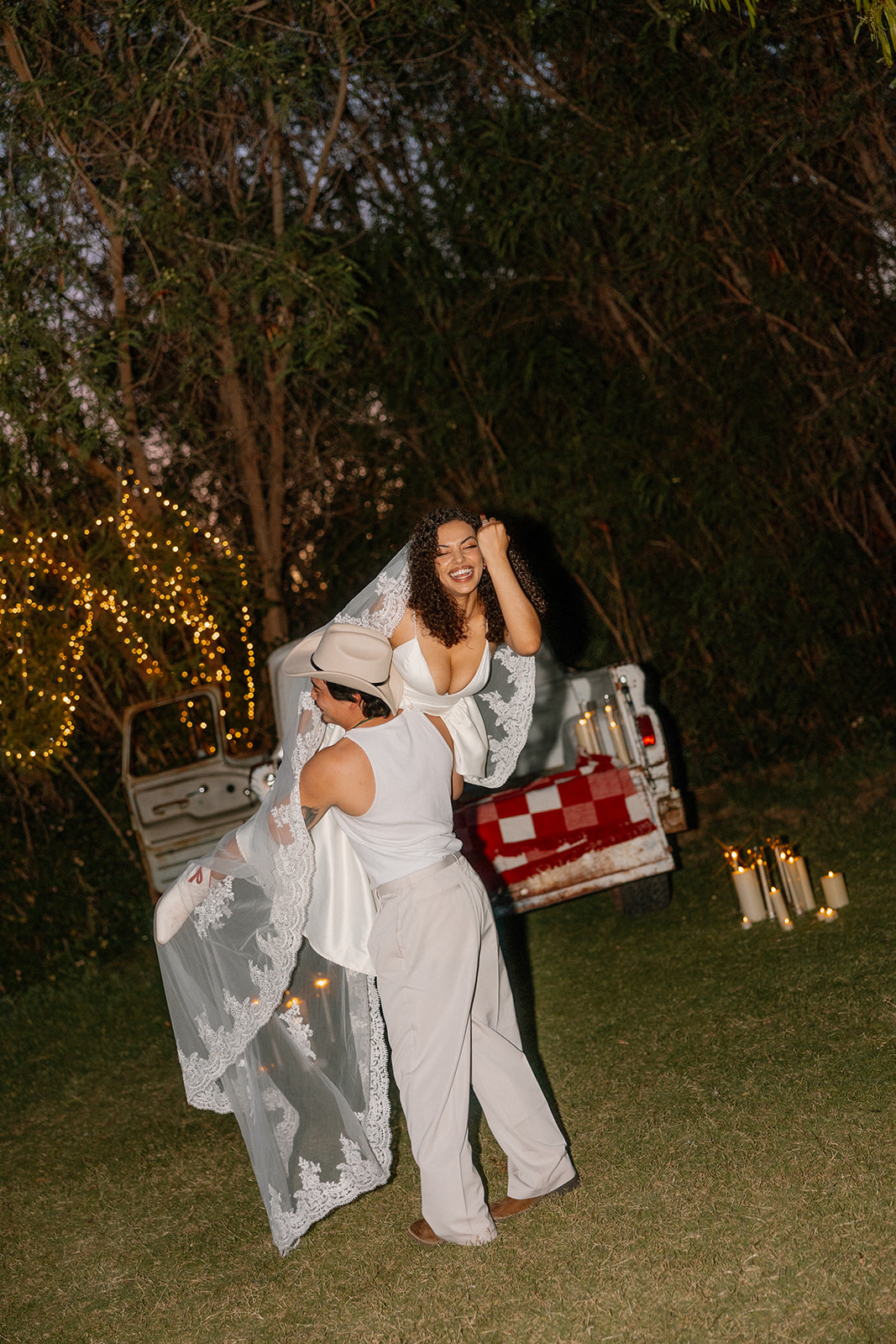 Captured mid-laugh, the groom lifts his bride as her lace veil flutters in the evening breeze—twinkle lights and a classic truck bed glowing in the background of this Western wedding scene.