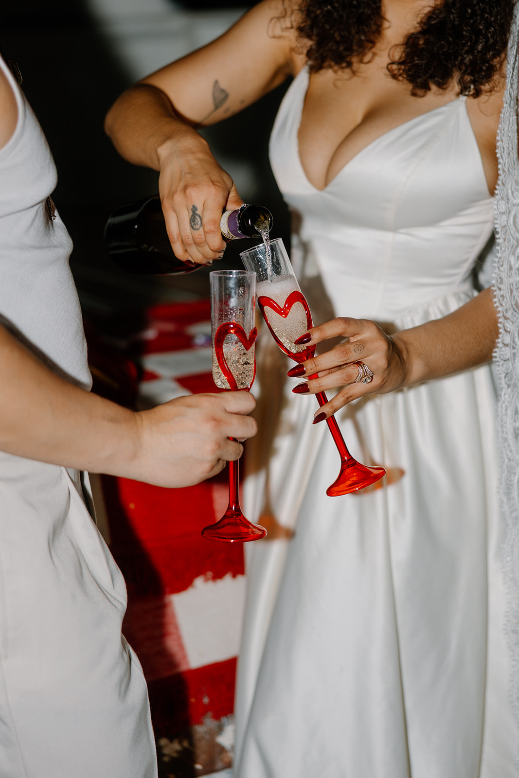 Close-up of the couple clinking champagne flutes with red heart stems during their Western wedding celebration. The bride wears a satin dress, and the groom sports a tank and slacks for a modern rustic look.