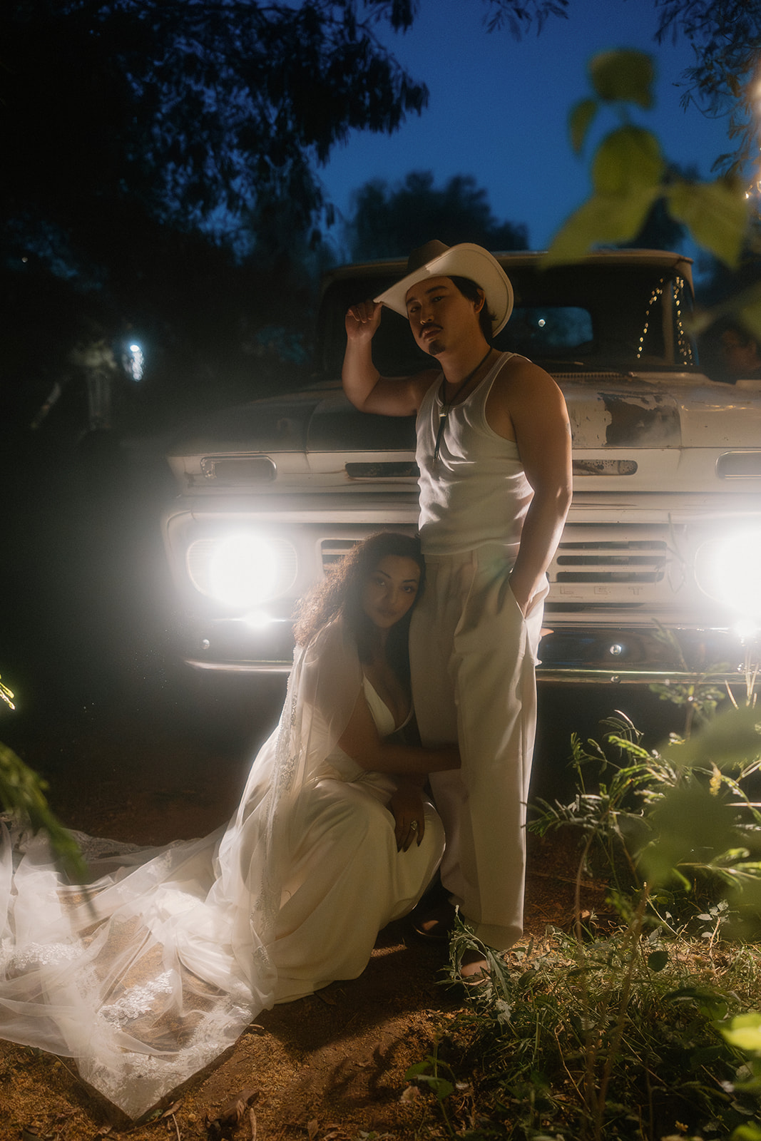 A bride in a white dress and veil kneels beside an old white pickup truck at night, embracing the leg of her groom who stands beside her in a cowboy hat and tank top. The truck's headlights are on, casting a dramatic glow in the evening setting.