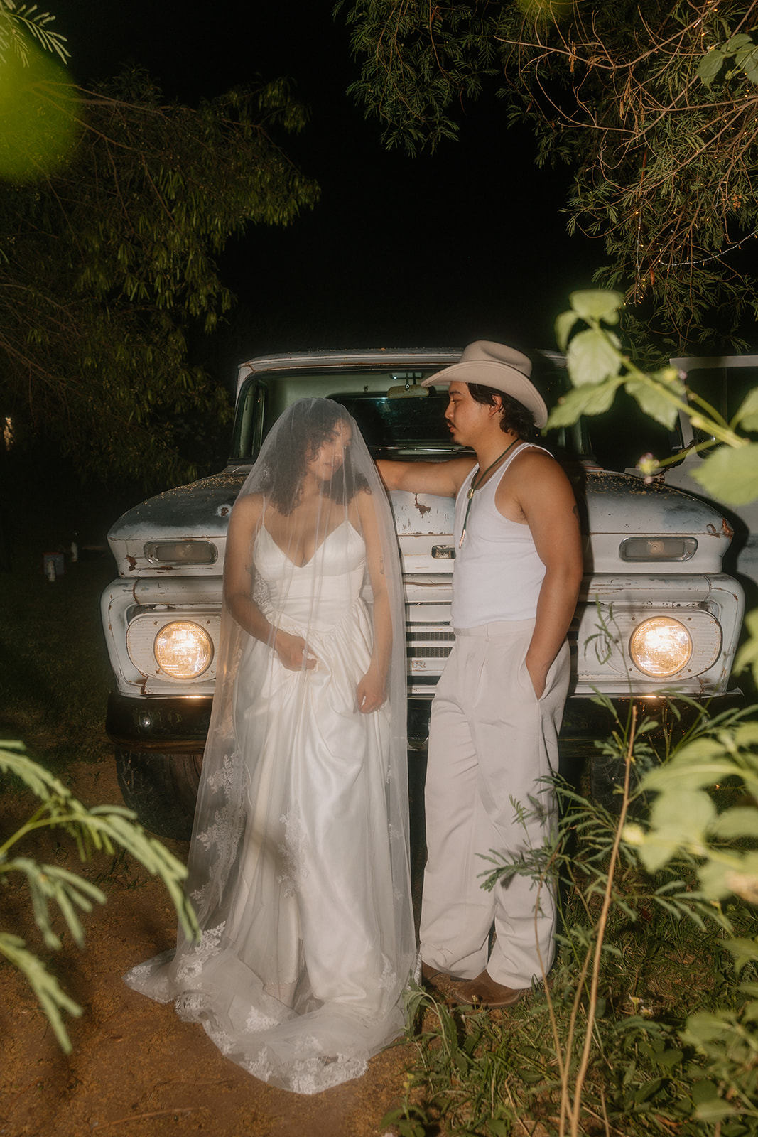 A nighttime portrait of the couple standing in front of the truck. The bride gazes at the groom through her veil, while he leans casually against the truck, both framed by greenery and headlights.